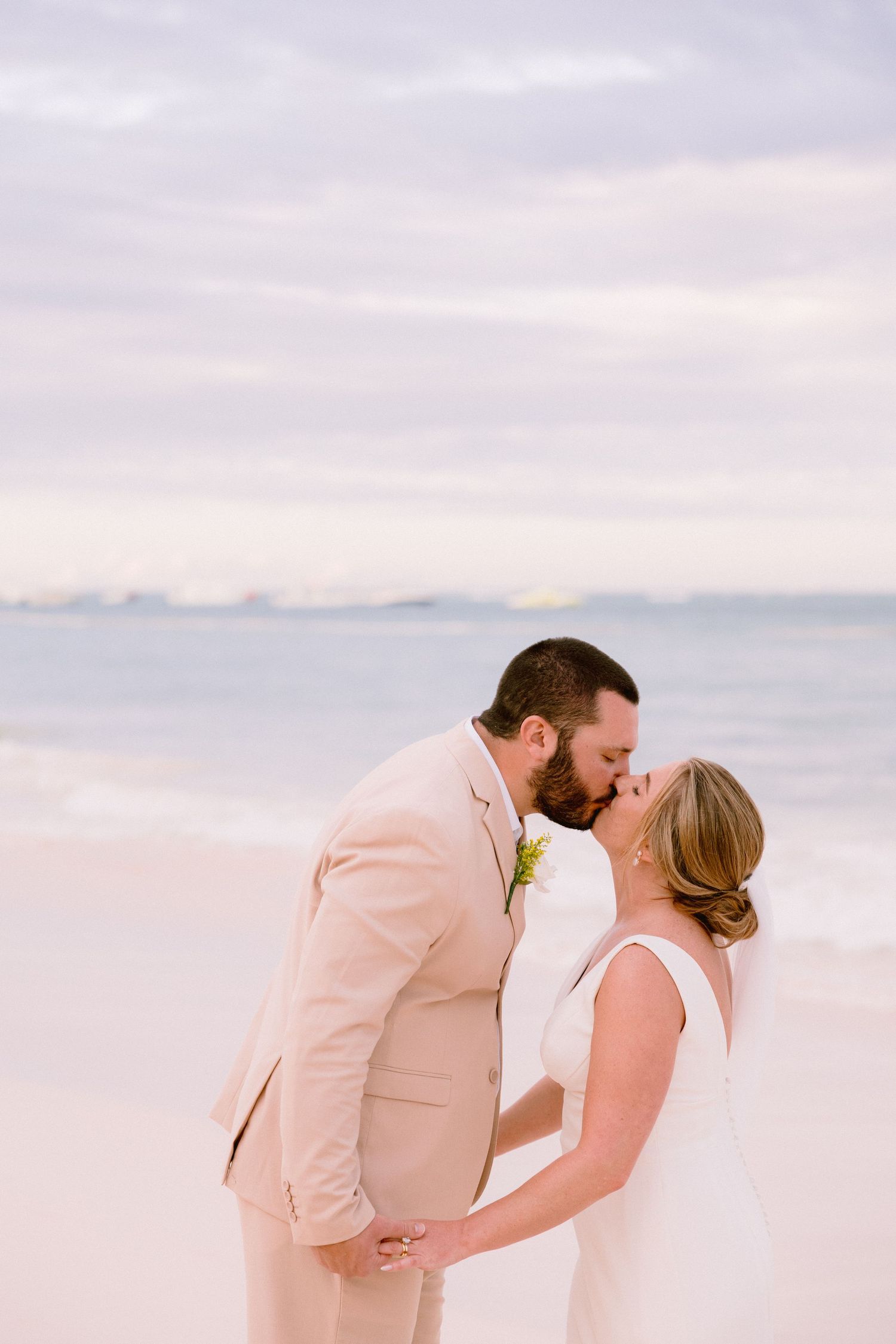 Bride and groom share a romantic kiss on a beach at sunset, with the groom in a blush suit and bride in a white gown.
