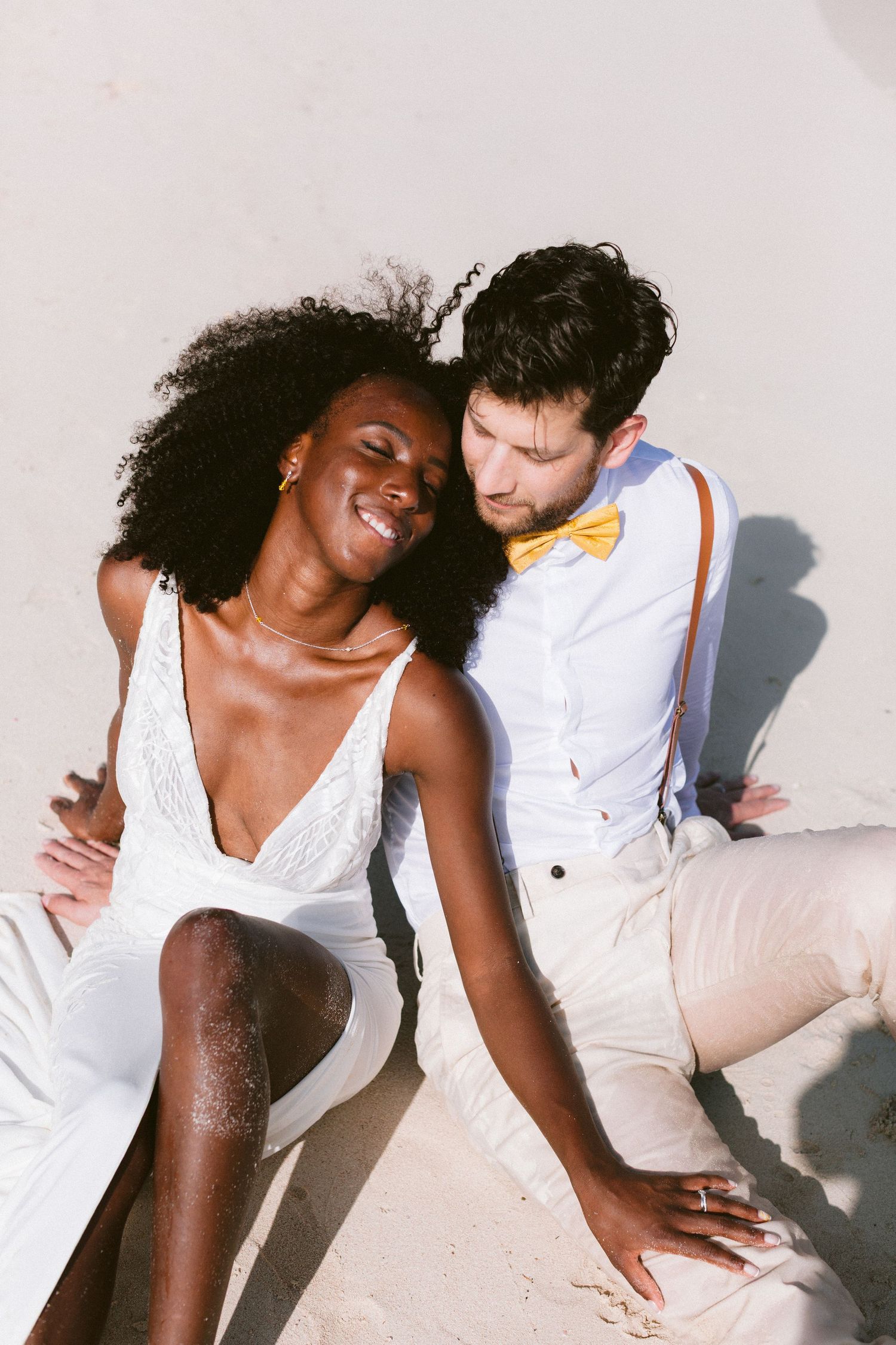 Interracial couple in wedding attire sitting on white sand beach, bride in white dress, groom in bow tie.