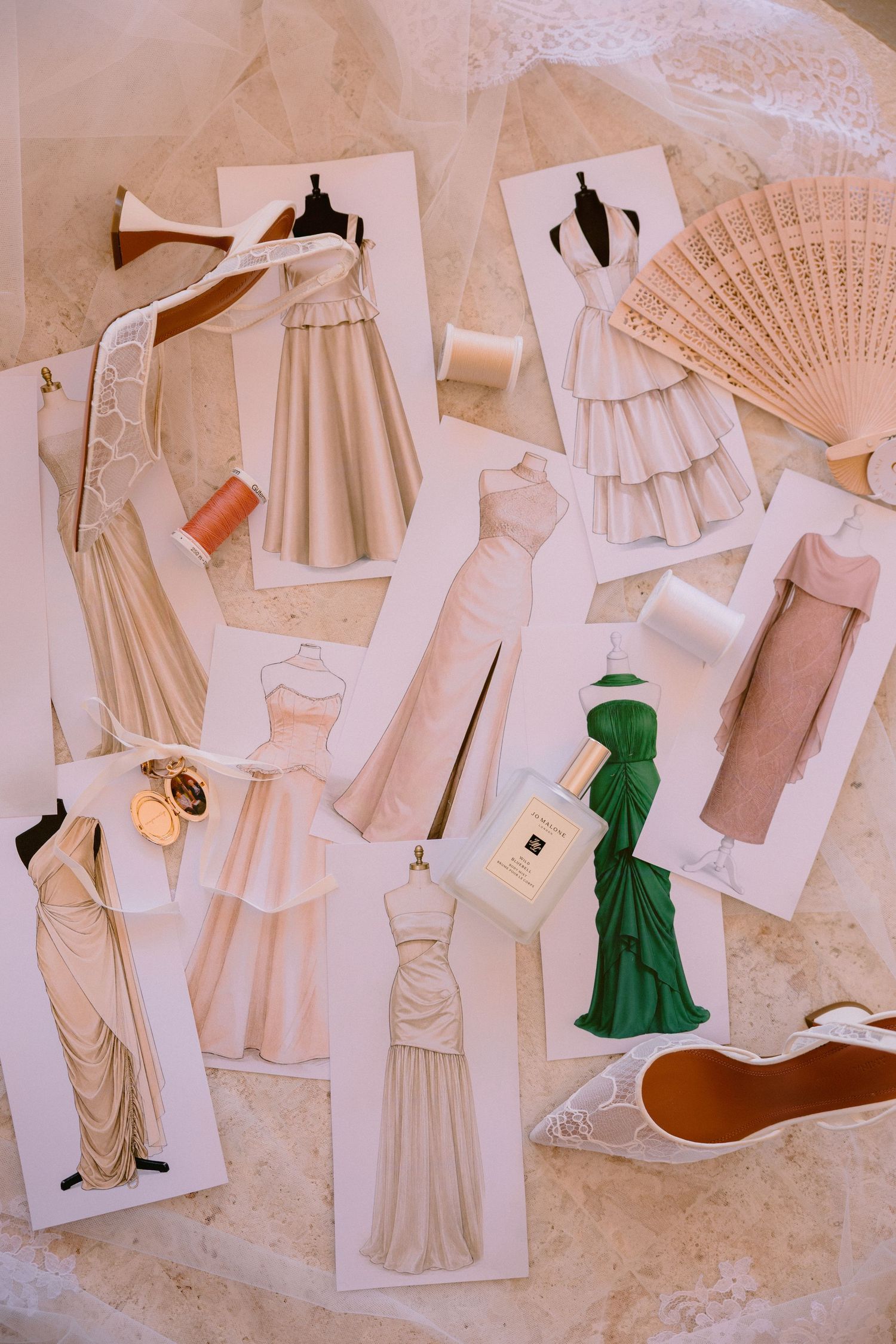 Fashion design sketches of elegant gowns in blush and green tones, displayed with spools of thread, perfume, and a lace shoe.