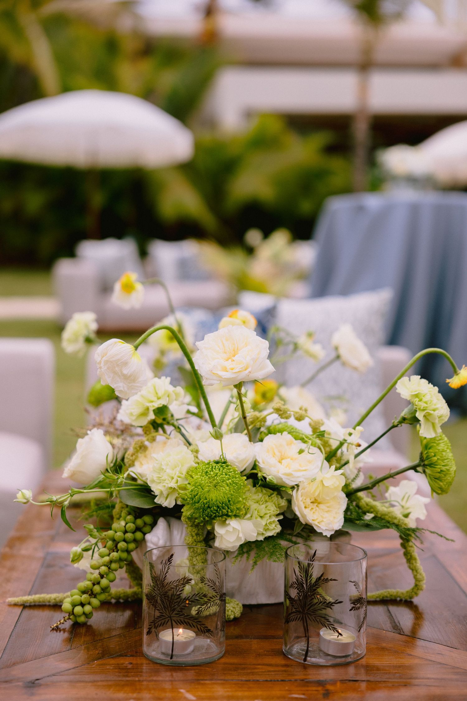 White and green floral centerpiece with roses and hydrangea on a rustic wooden table, with glass candle holders at an outdoor event.