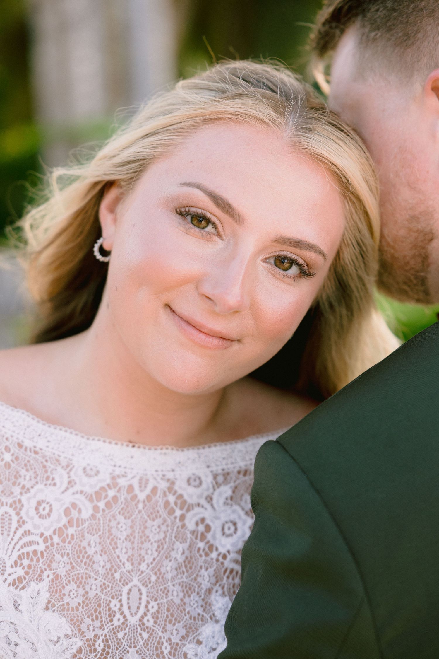 Smiling blonde bride in white lace dress with pearl earrings, leaning against groom in green suit outdoors.