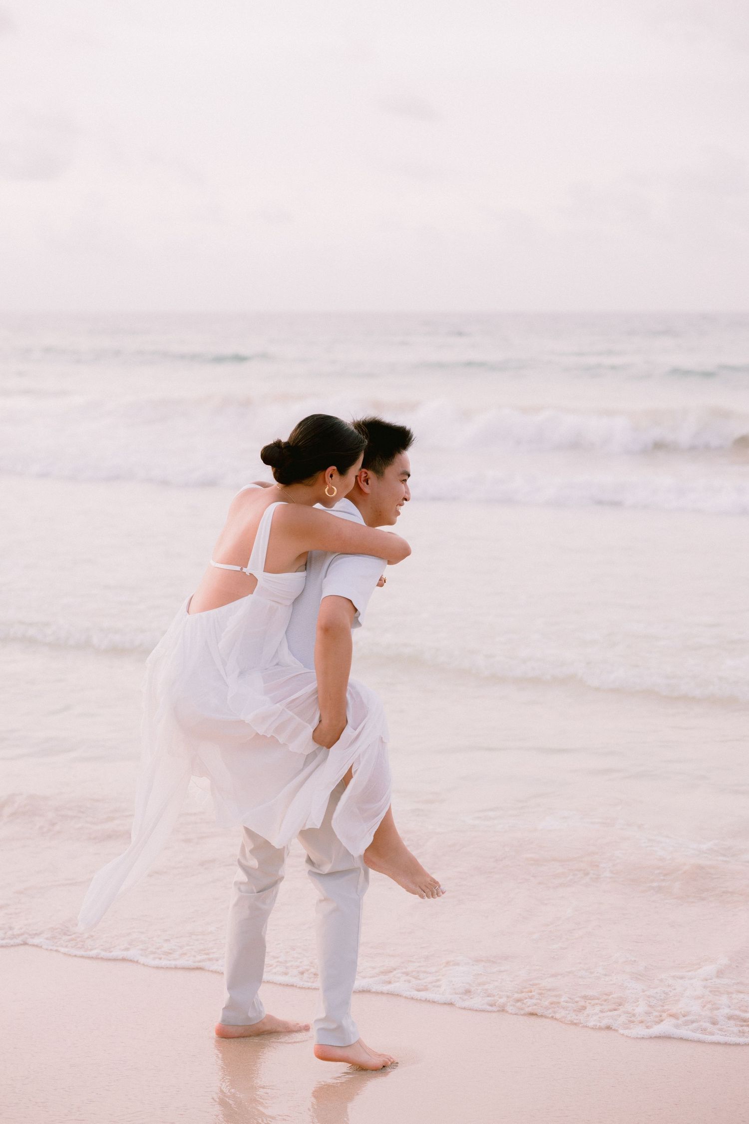Couple in white attire sharing a romantic piggyback ride on a serene beach with gentle ocean waves.