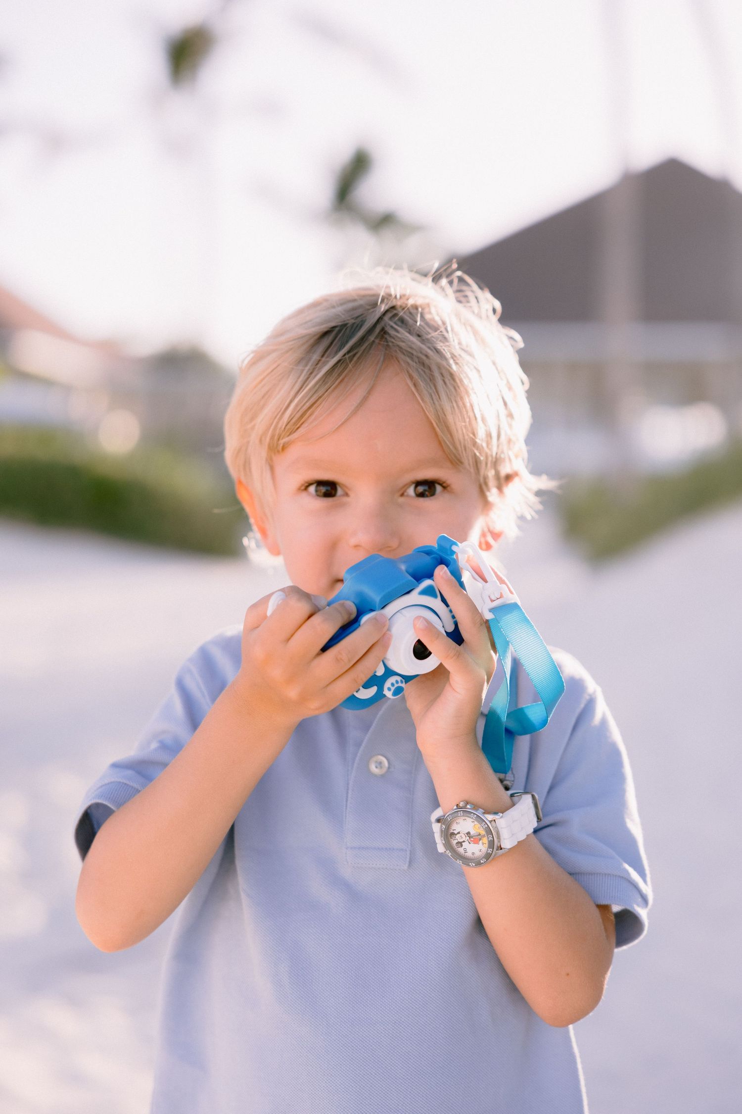 Young blond boy in blue polo shirt holding a toy camera up to his face outdoors in bright sunlight.
