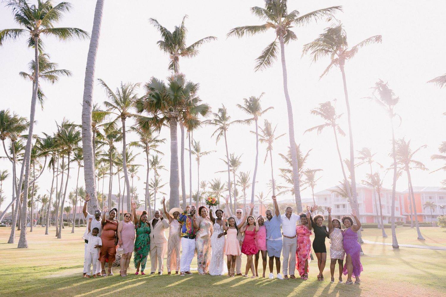 A large group of wedding guests pose together on a tropical beach with palm trees at sunset.