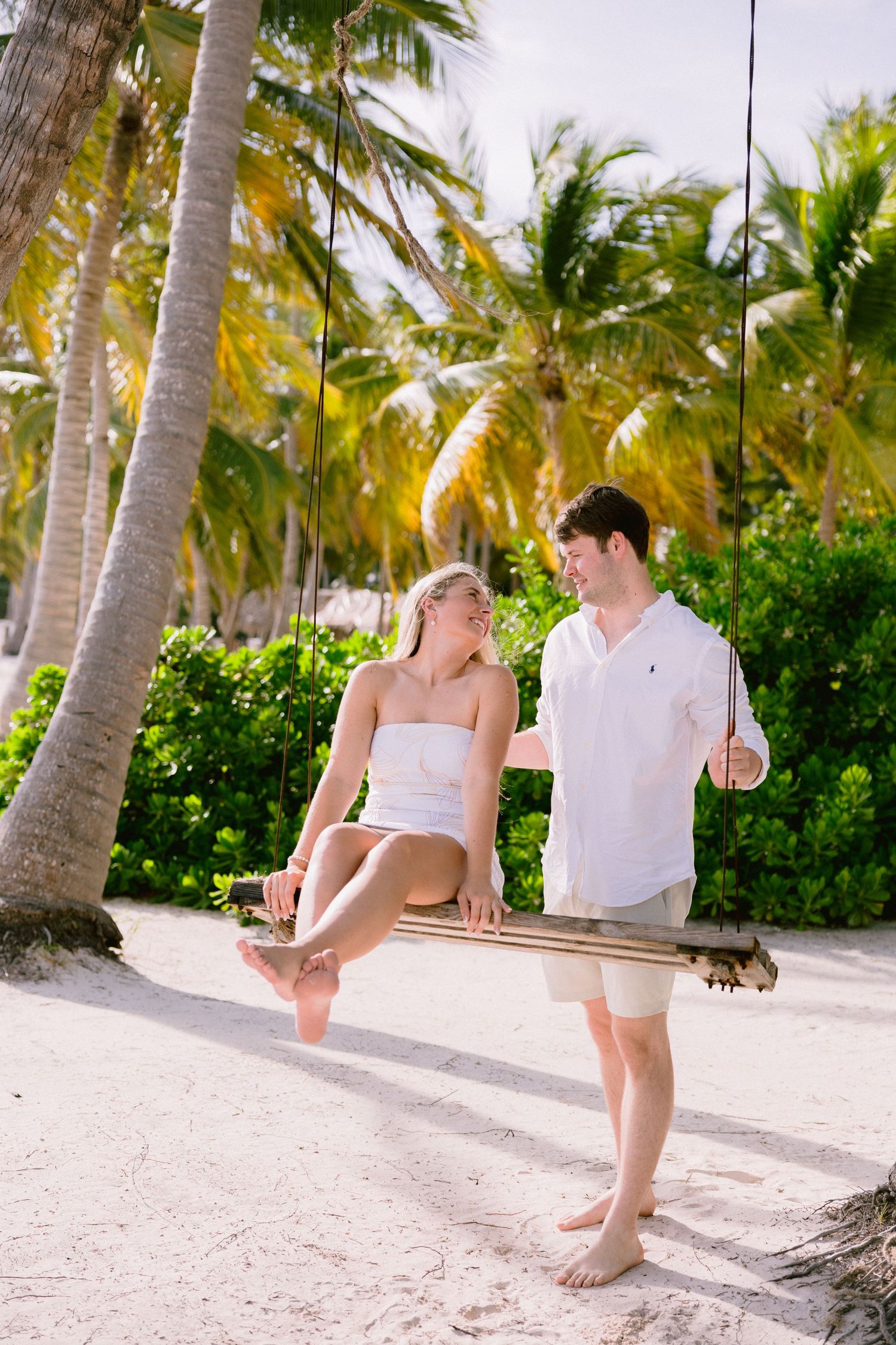 A couple sits together on a palm tree swing at a tropical beach with white sand and lush greenery.