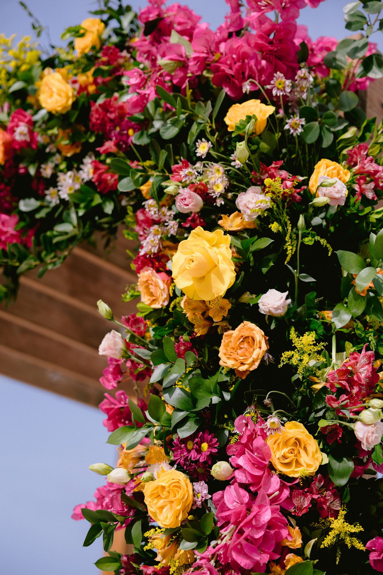 A vibrant floral arrangement featuring pink, yellow, and peach roses with lush green foliage against a blue background.