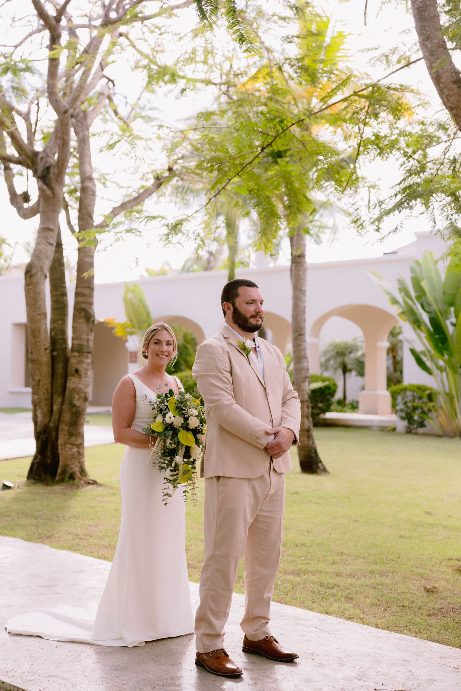 Bride in white gown holding bouquet stands with groom in tan suit at outdoor wedding ceremony surrounded by tropical trees.