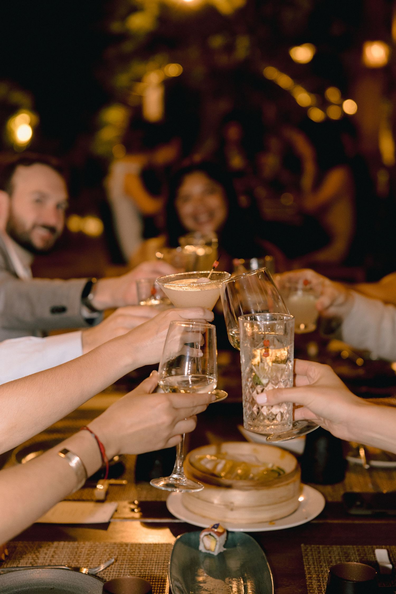 Friends raise cocktails and glasses in a lively toast over a candlelit restaurant table with a bamboo steamer of dumplings.