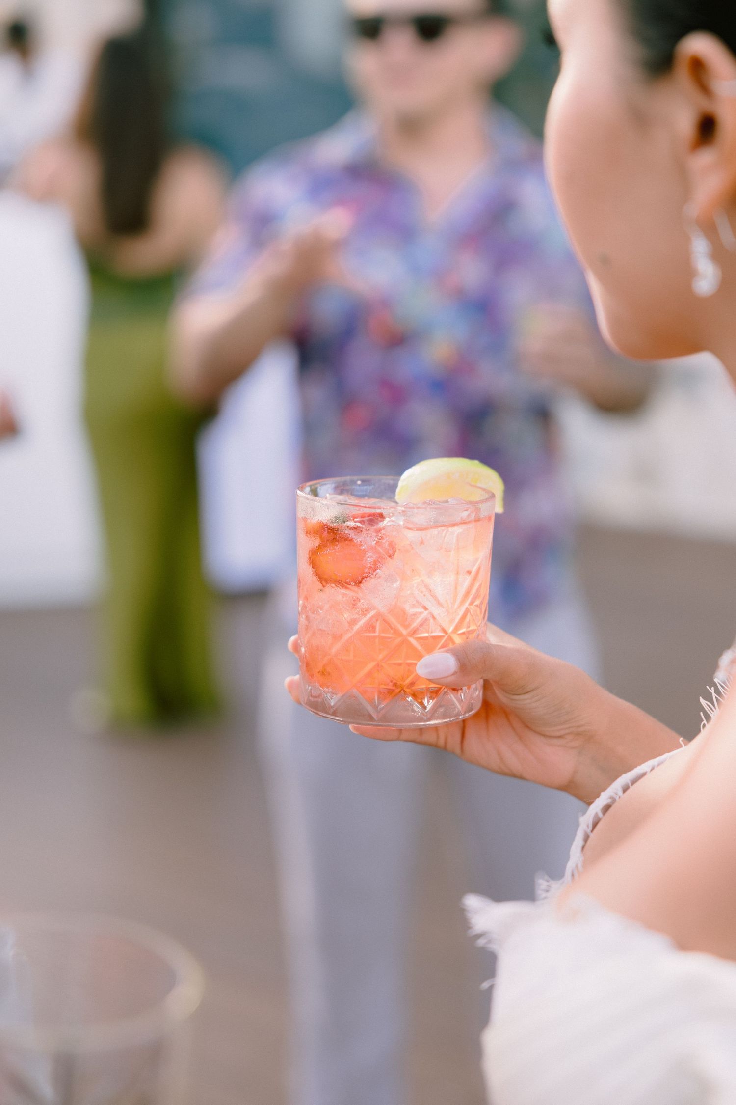 Woman in white holds a pink strawberry cocktail with a lime wheel at an outdoor party while guests mingle in the blurred background.