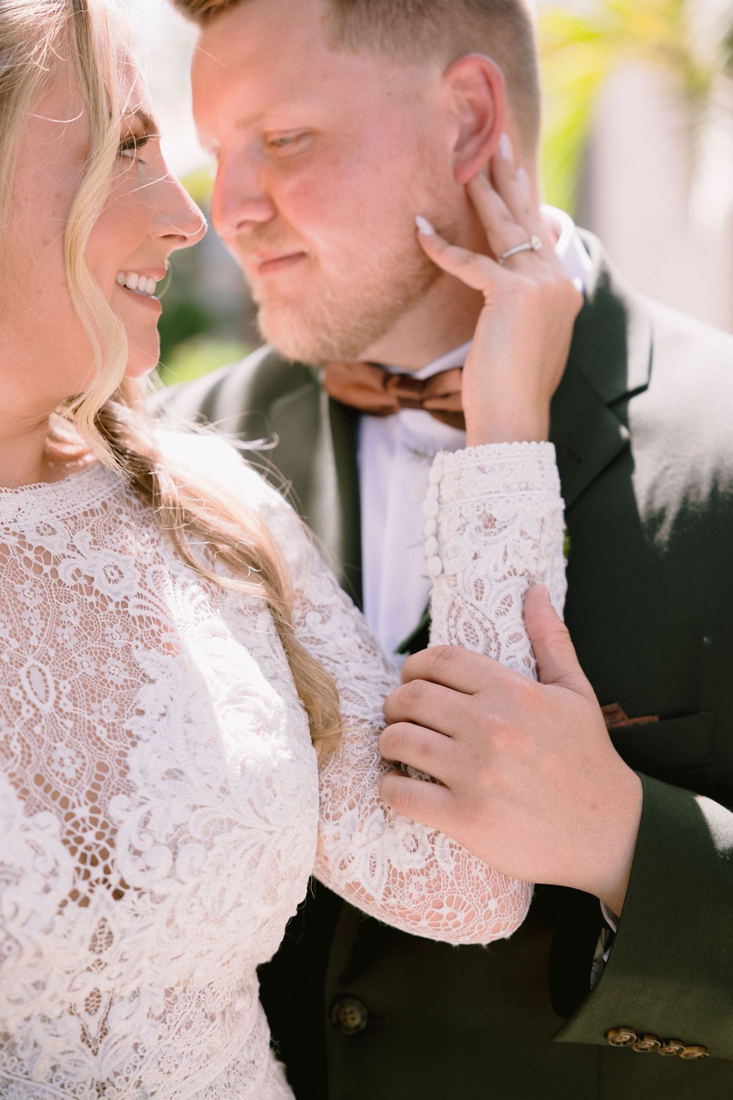 Bride in lace gown tenderly touches groom's face as he leans in, both wearing wedding rings outdoors.