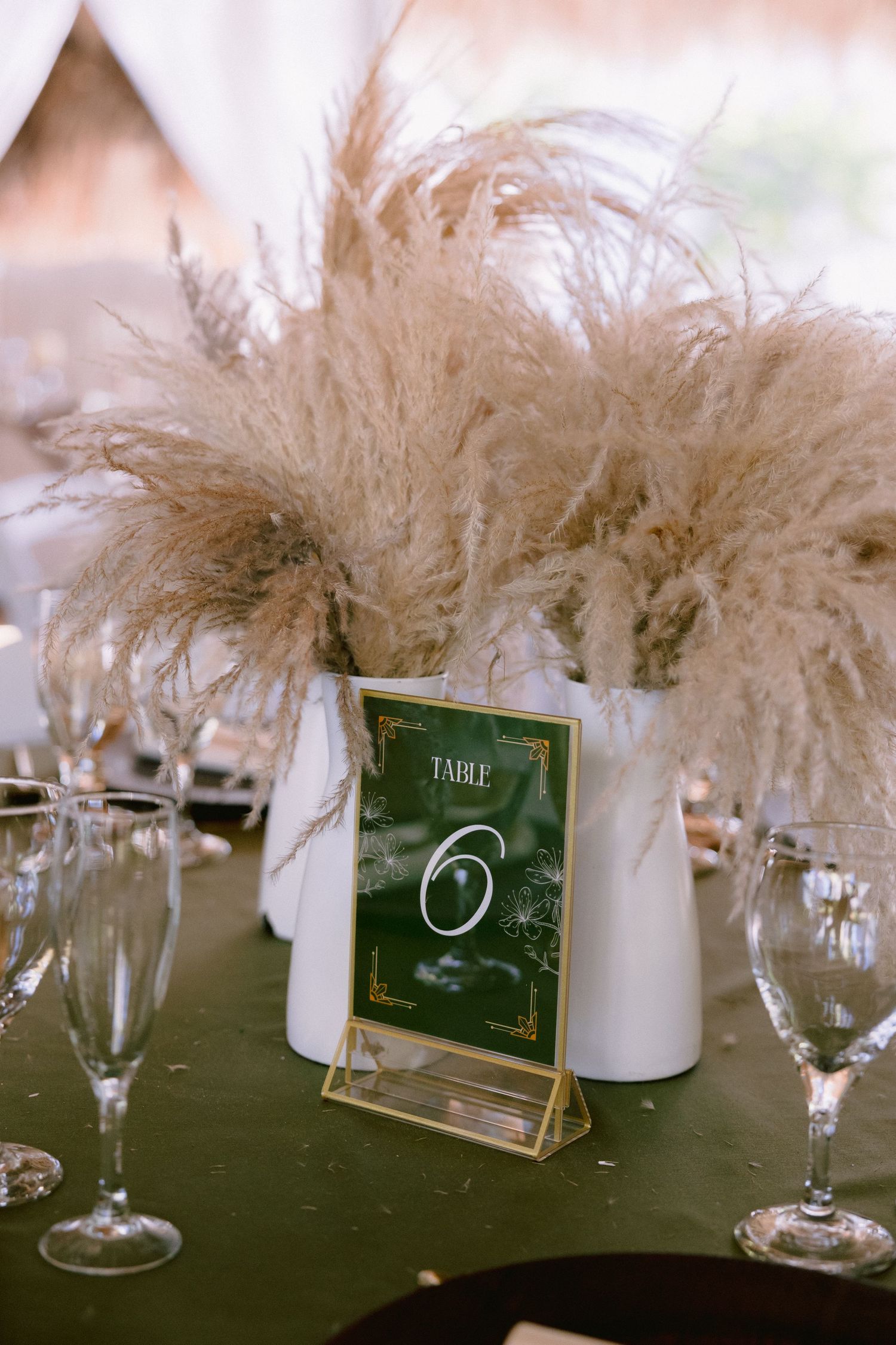 Wedding table 6 centerpiece with pampas grass in white vases on green tablecloth with gold-framed table number sign.