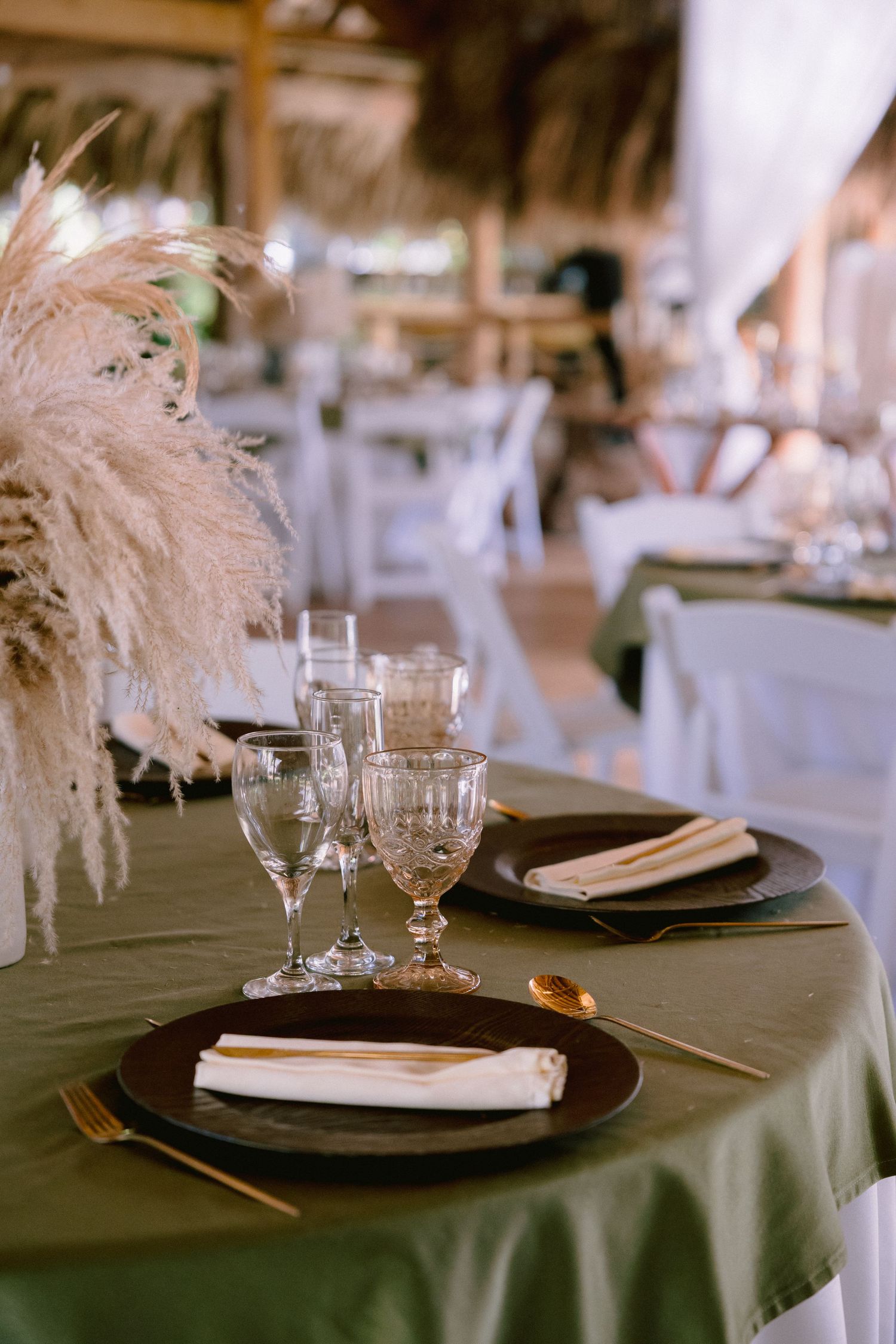 Elegant wedding table setting with green tablecloth, dark plates, gold cutlery, crystal glasses, and pampas grass centerpiece.