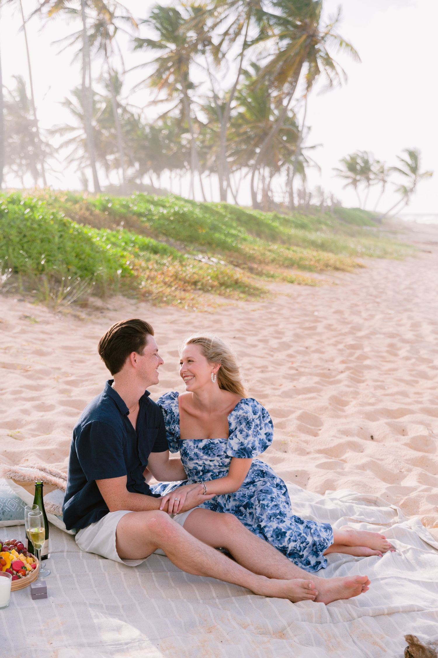 A couple enjoys a romantic picnic on a tropical beach with palm trees swaying in the background.