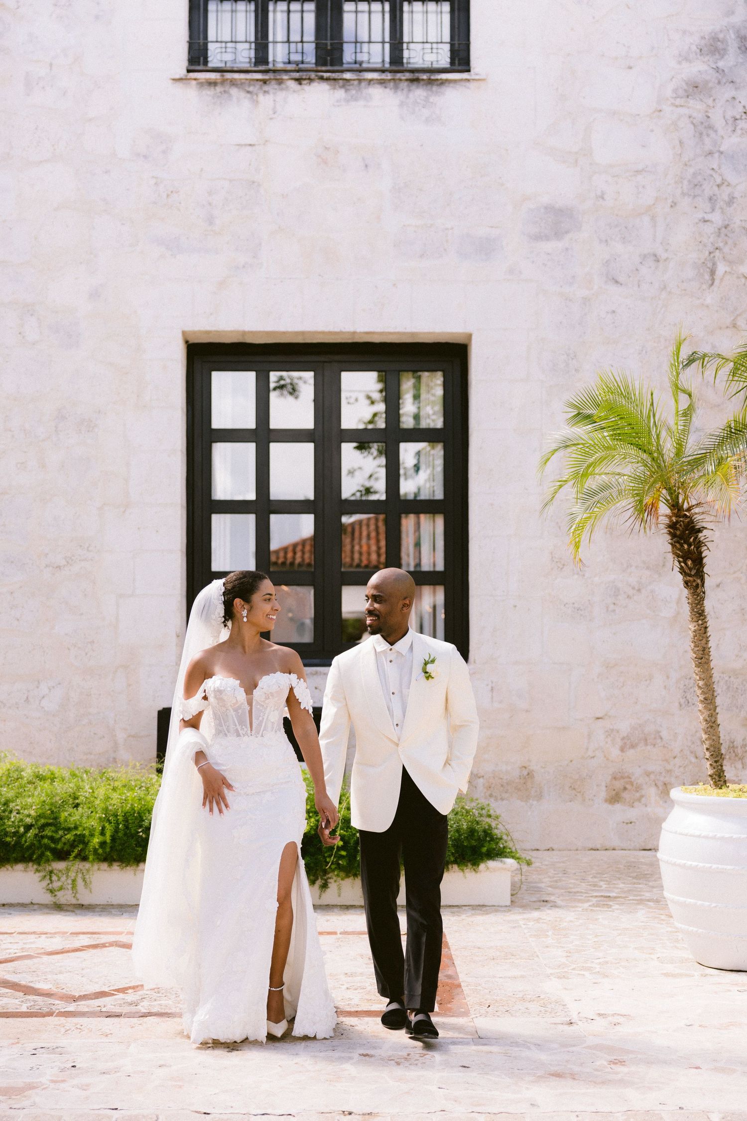 A couple in elegant wedding attire walks together outside a white stucco building with large windows and a palm tree.
