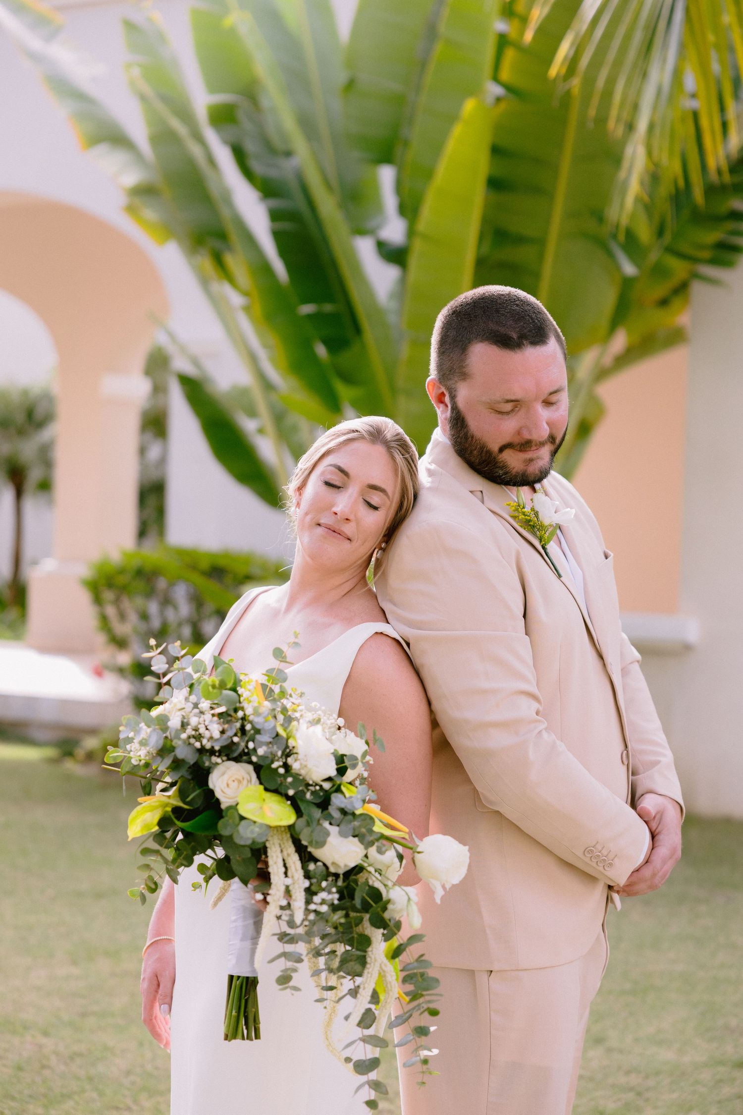 A romantic wedding couple embraces in front of tropical palm trees while wearing light beige and white attire.