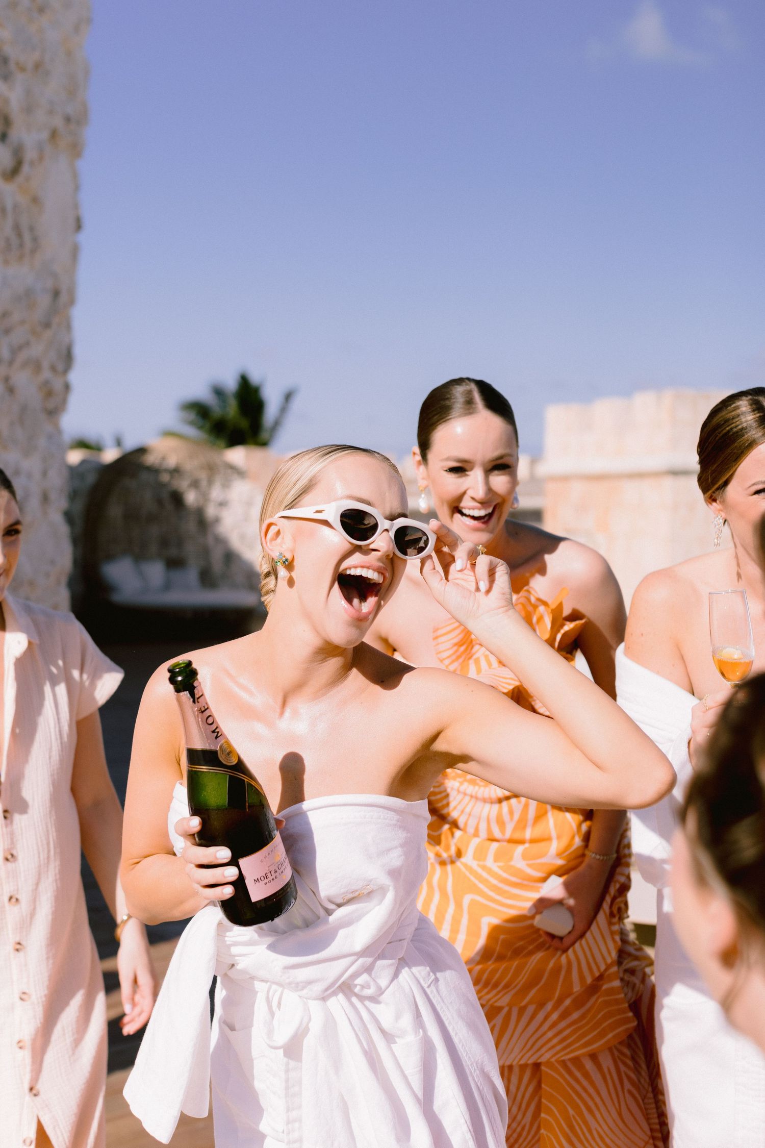 A group of bridesmaids in orange and white dresses laugh together during a wedding celebration outdoors.