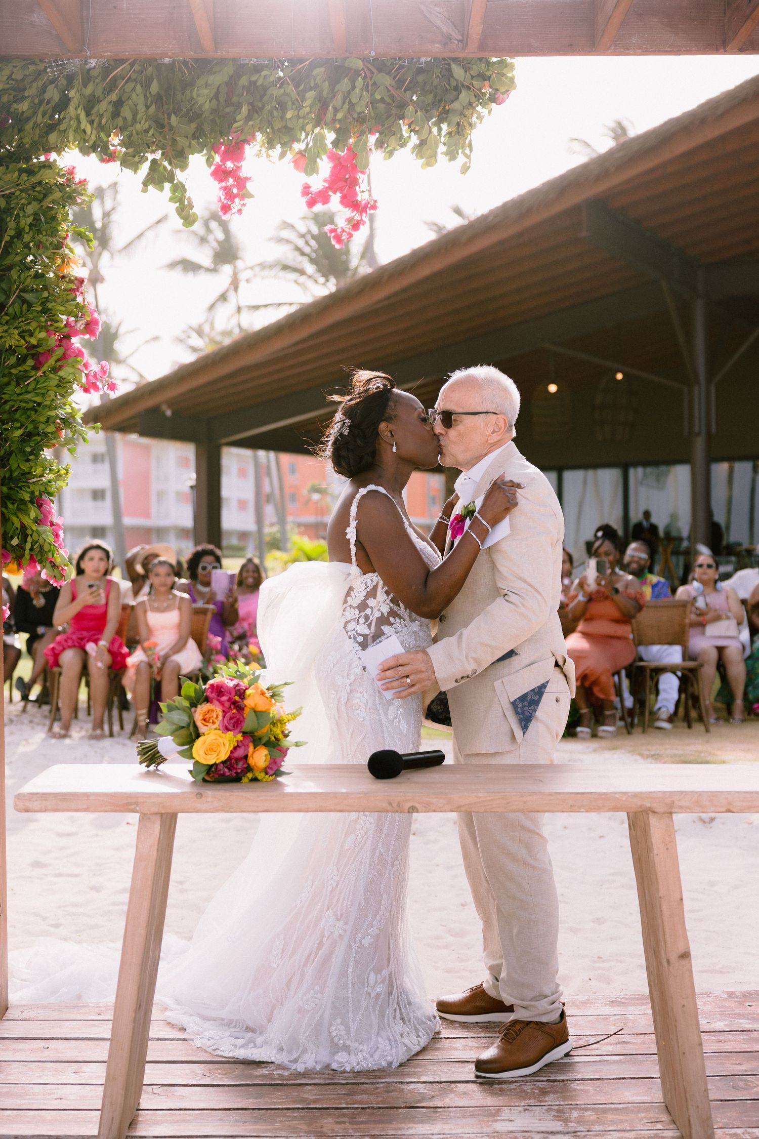 A romantic wedding ceremony takes place on a wooden deck with pink flowers and palm trees in the background.