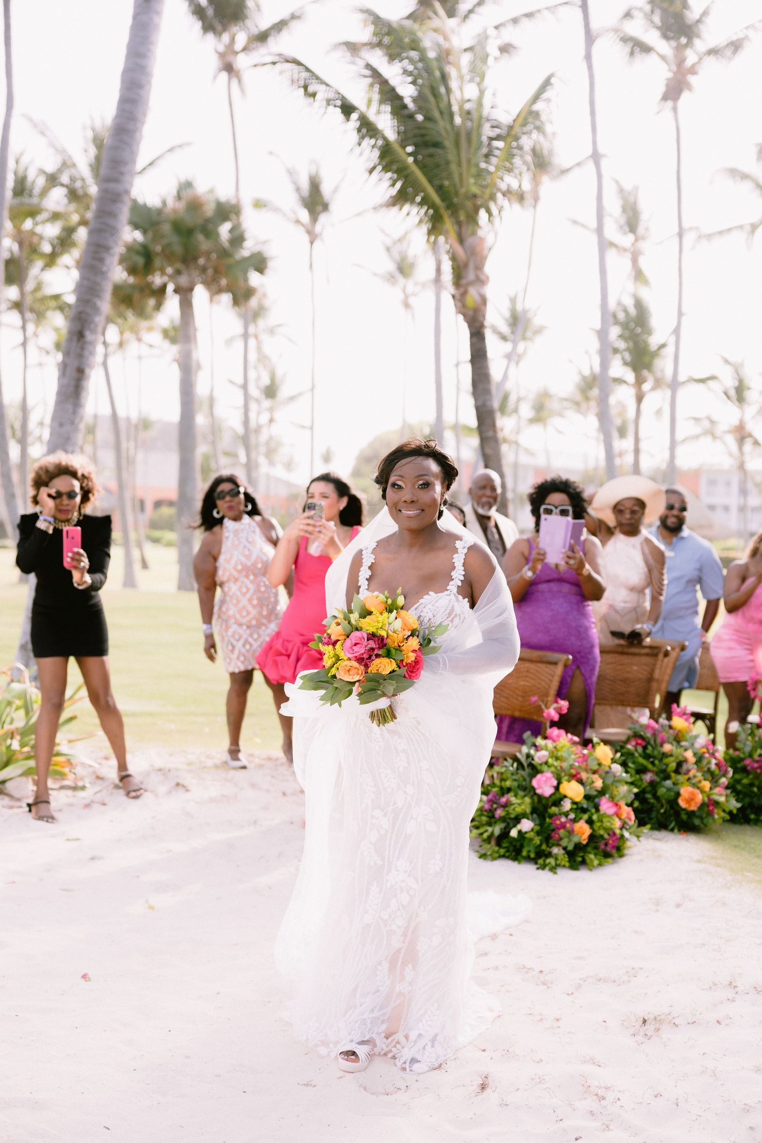 Tropical beach wedding ceremony setup with palm trees and floral arrangements along sandy pathway.