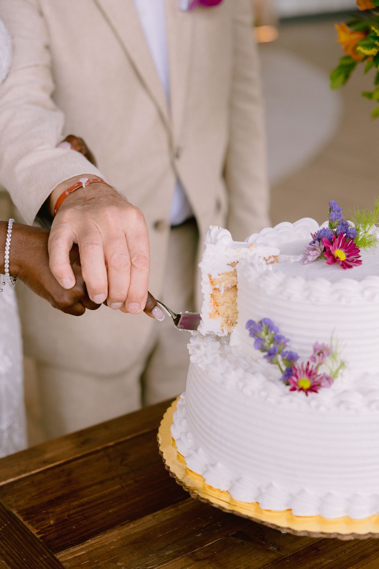 Hands cutting into a white frosted wedding cake decorated with fresh purple and pink flowers.