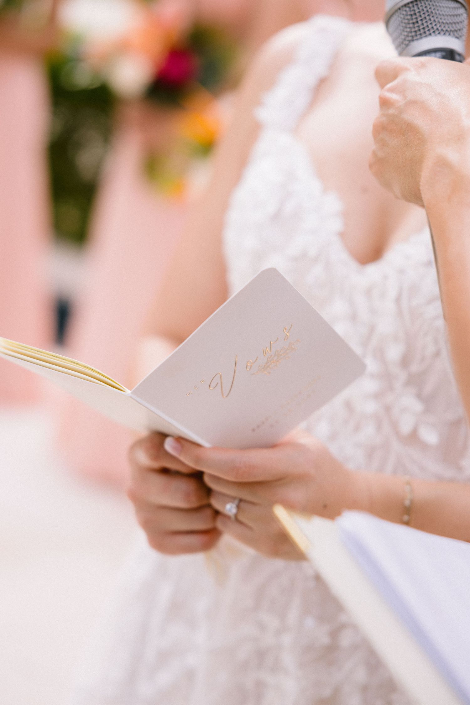 Close-up view of hands holding white wedding vows or ceremony papers against a soft pink floral background.