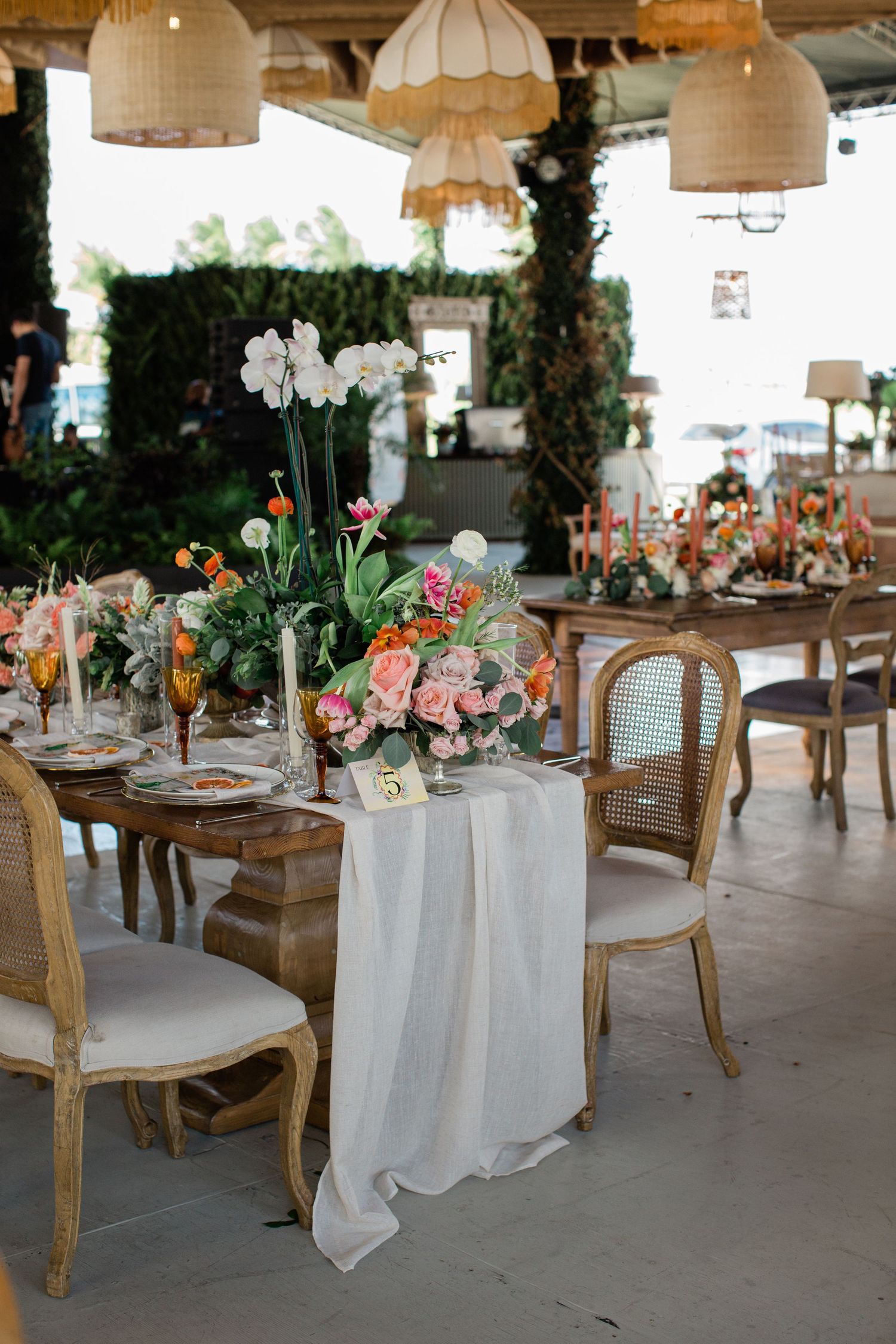 Elegant wedding reception tables with white linens, wooden chairs, and floral centerpieces under drum shade pendant lights.