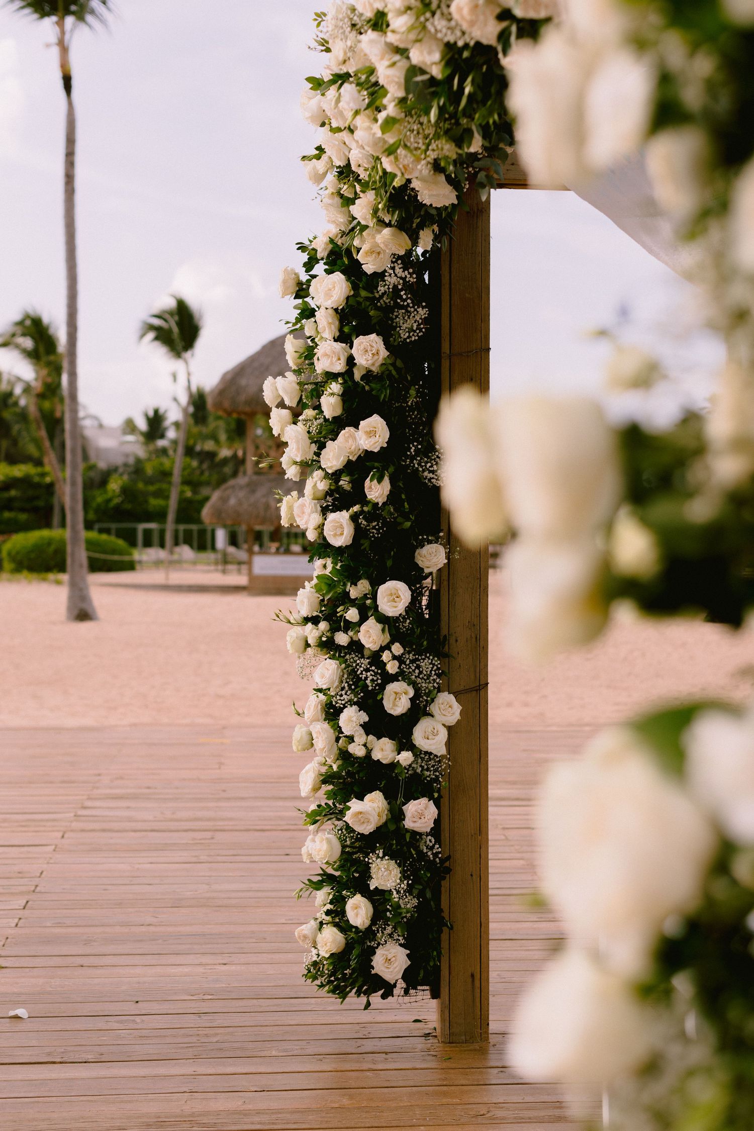 White roses adorn a wooden wedding arch on a tropical beach boardwalk with palm trees in the background.
