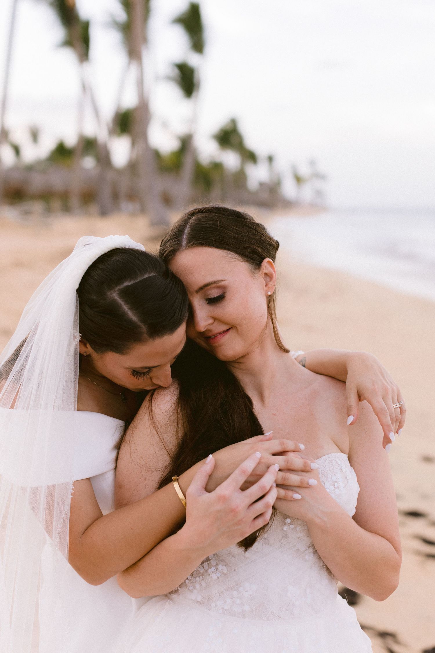 Two brides in white wedding dresses embrace tenderly on a tropical beach with palm trees in the background.