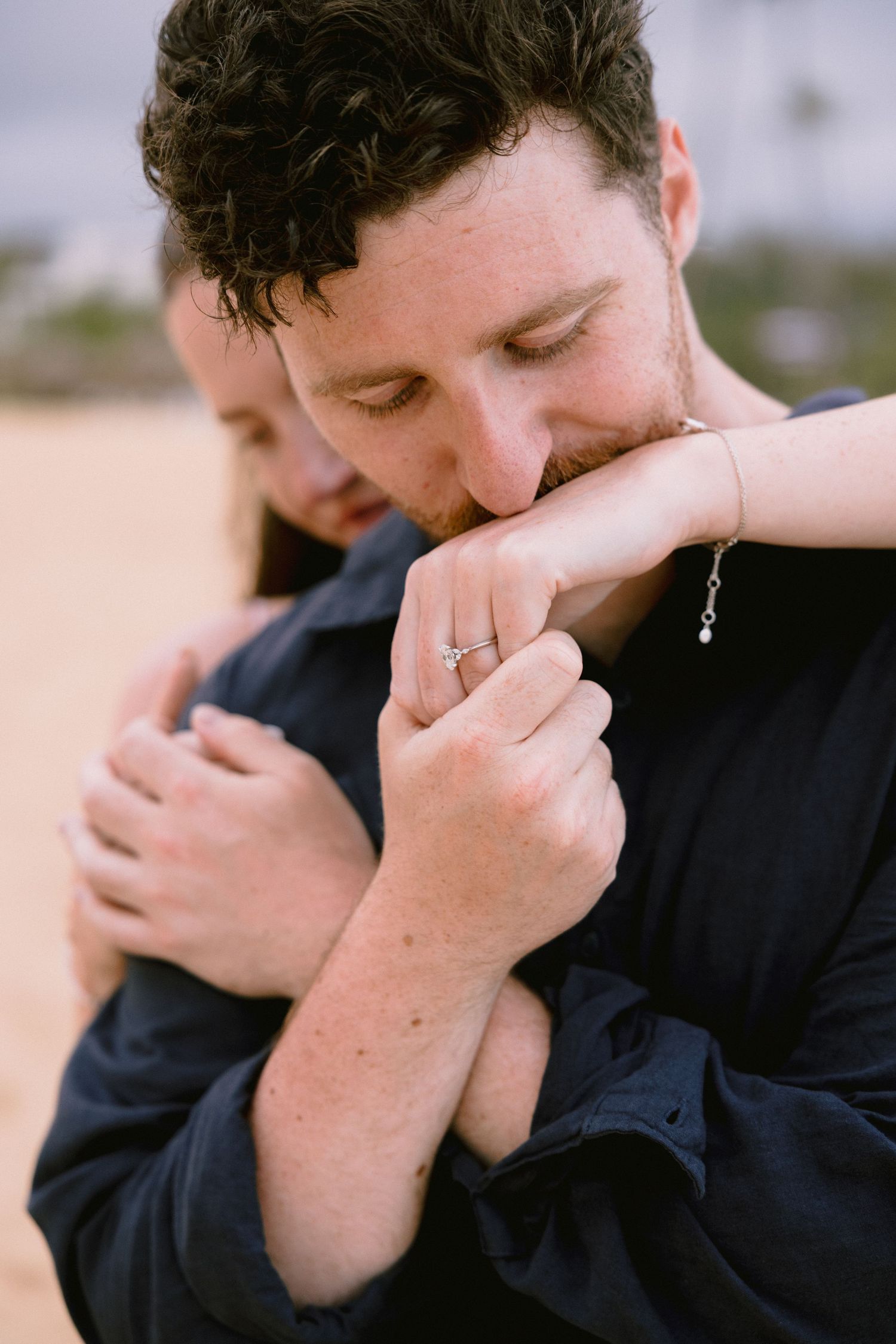 Man in navy shirt kissing woman's hand with engagement ring at sandy beach location.