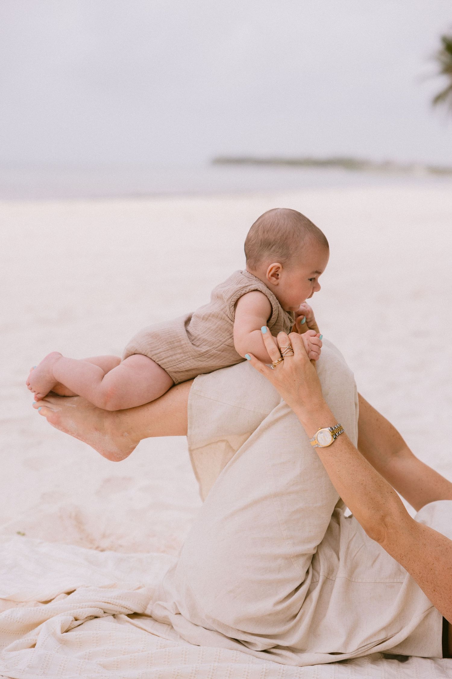 Baby in beige outfit playing on blanket at beach with palm trees.