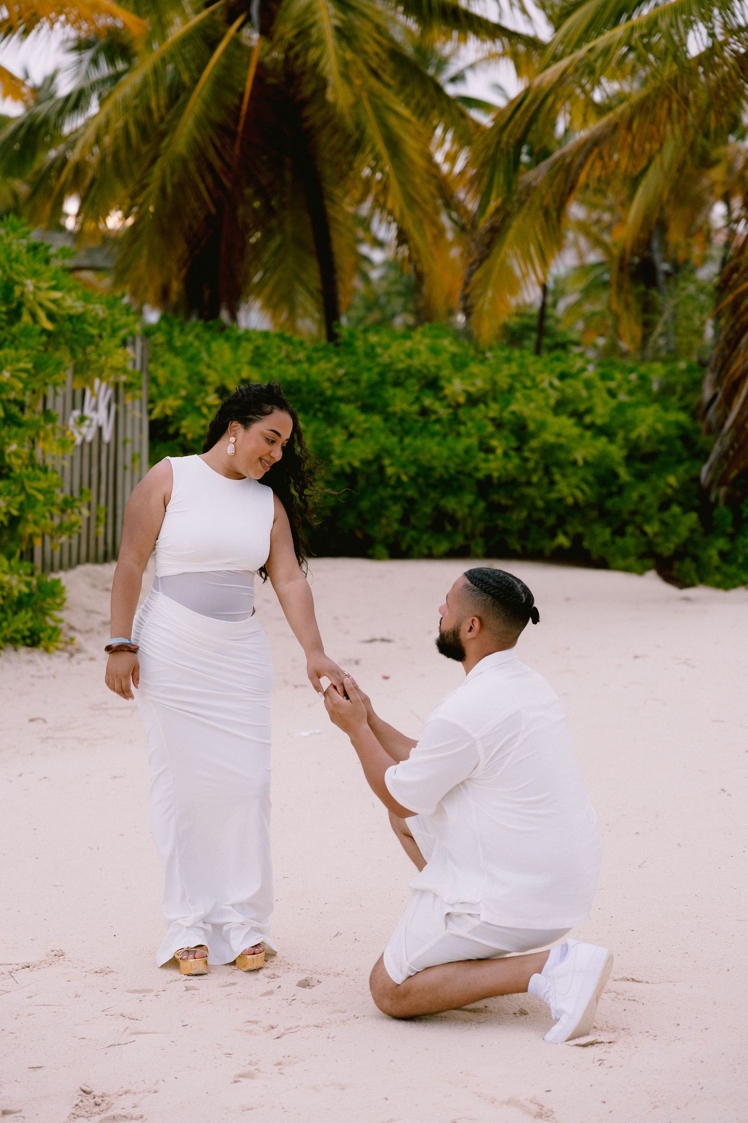 Couple in white outfits playfully posing on tropical beach with palm trees.