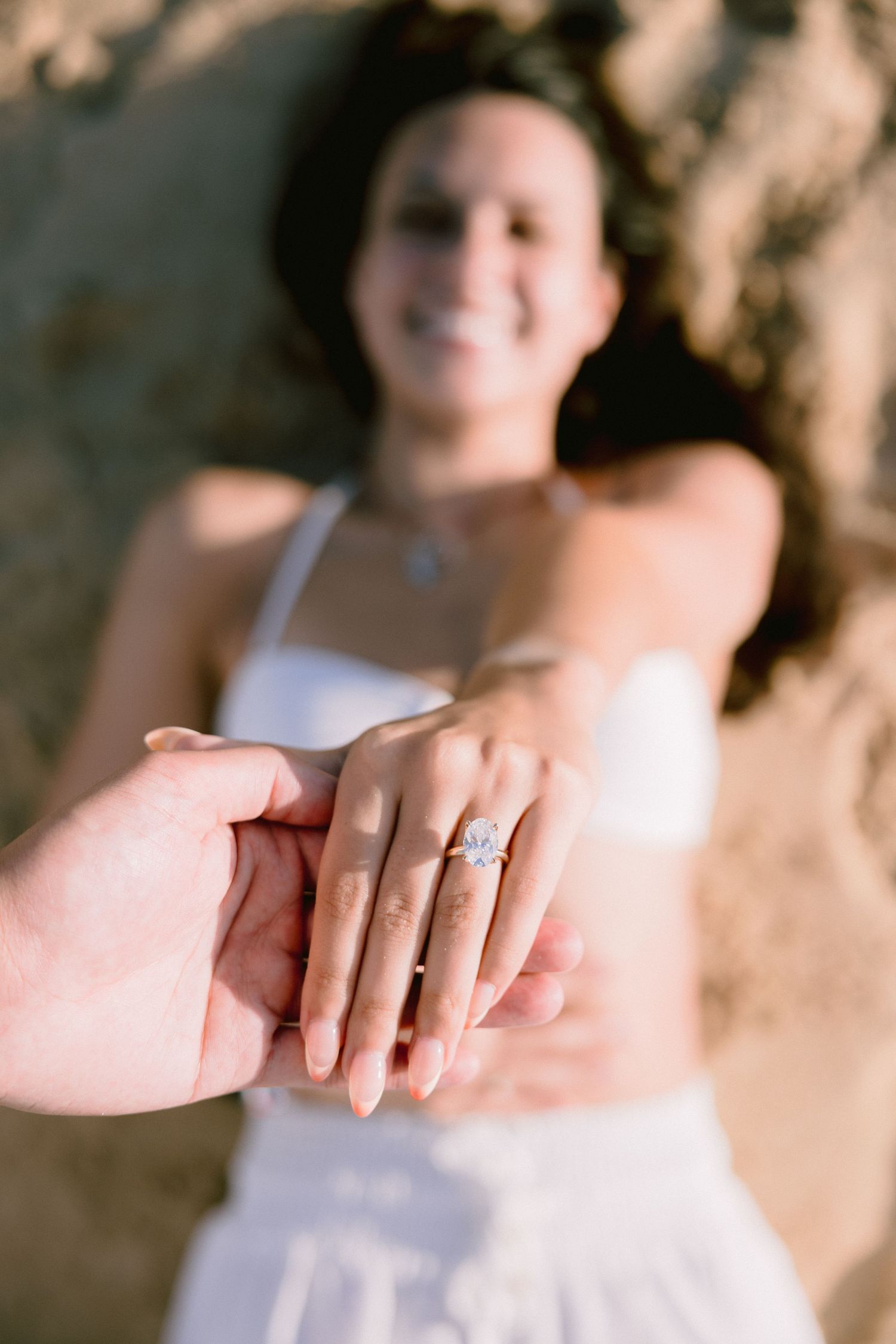 A smiling bride lying on sand displays her diamond engagement ring while holding hands with her partner.