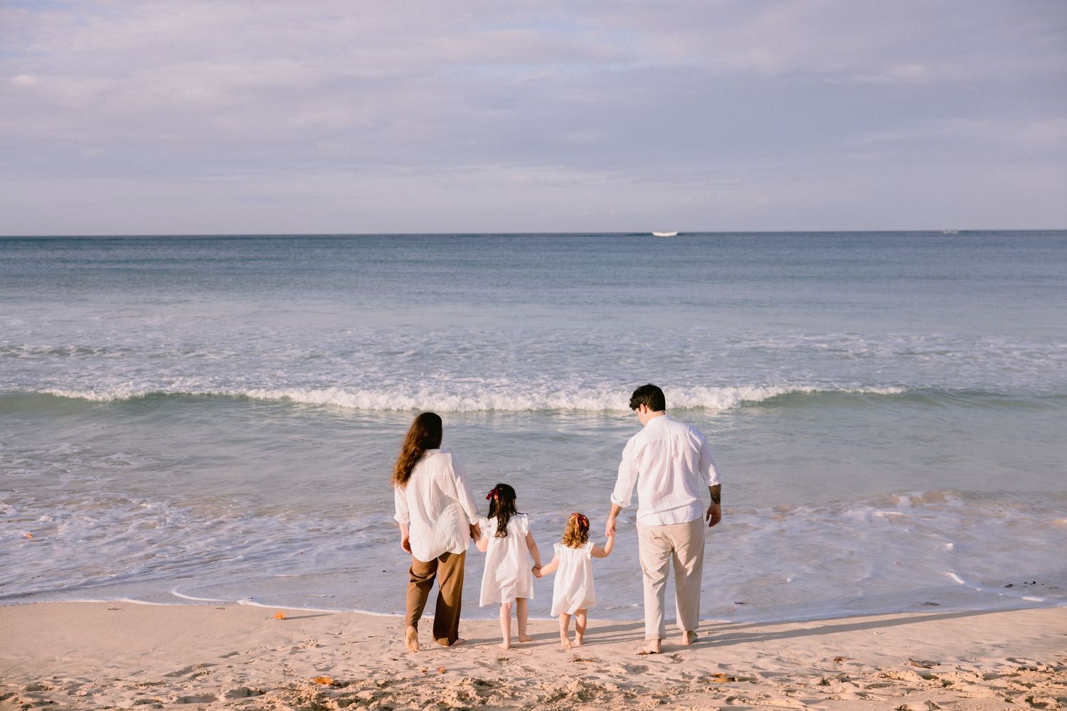 Family of four in white outfits holding hands on a sandy beach, gazing at ocean waves under a cloudy sky.