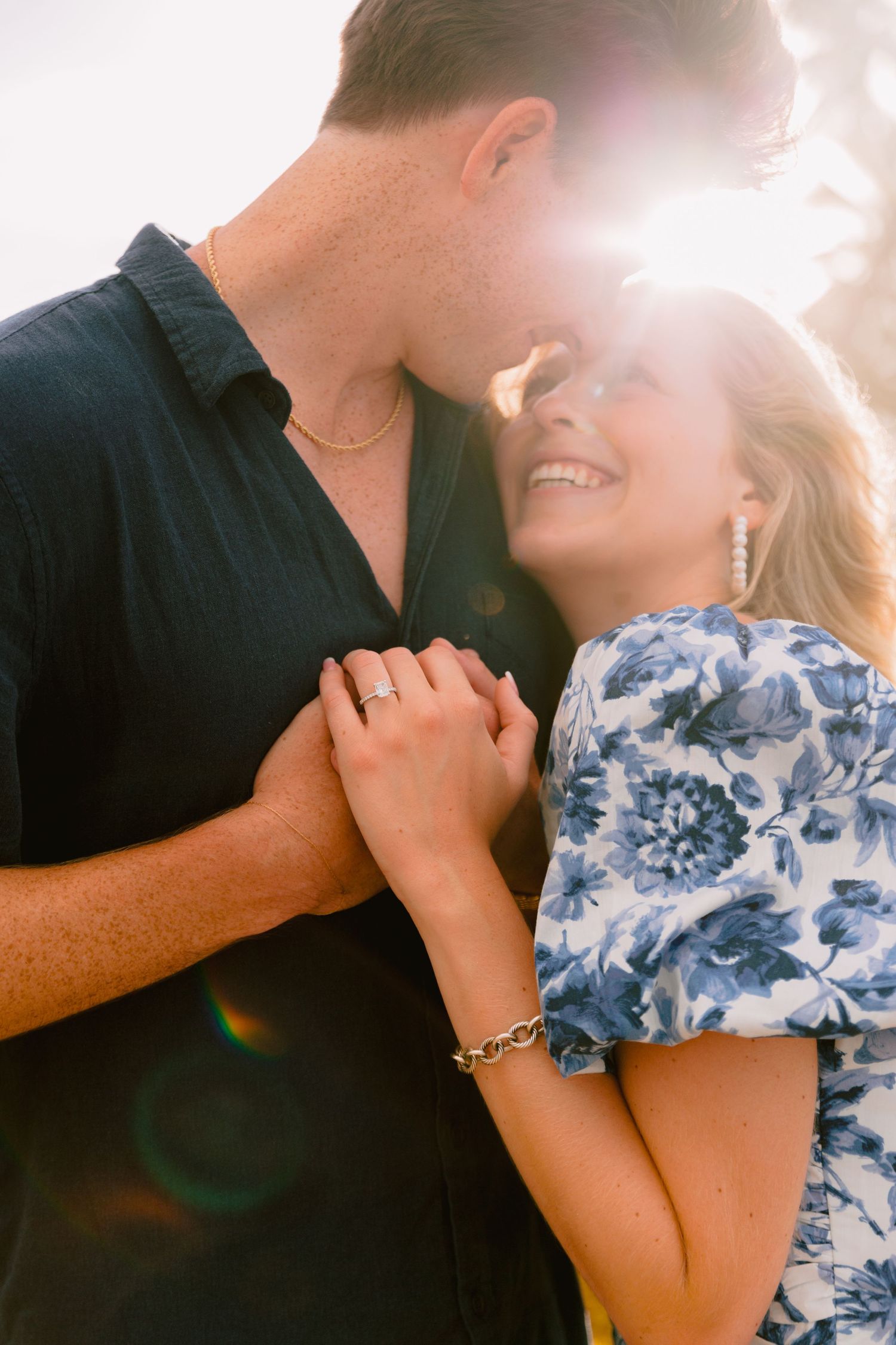 Romantic couple embraces in warm sunlight, with one wearing a blue floral dress and the other in a dark shirt.