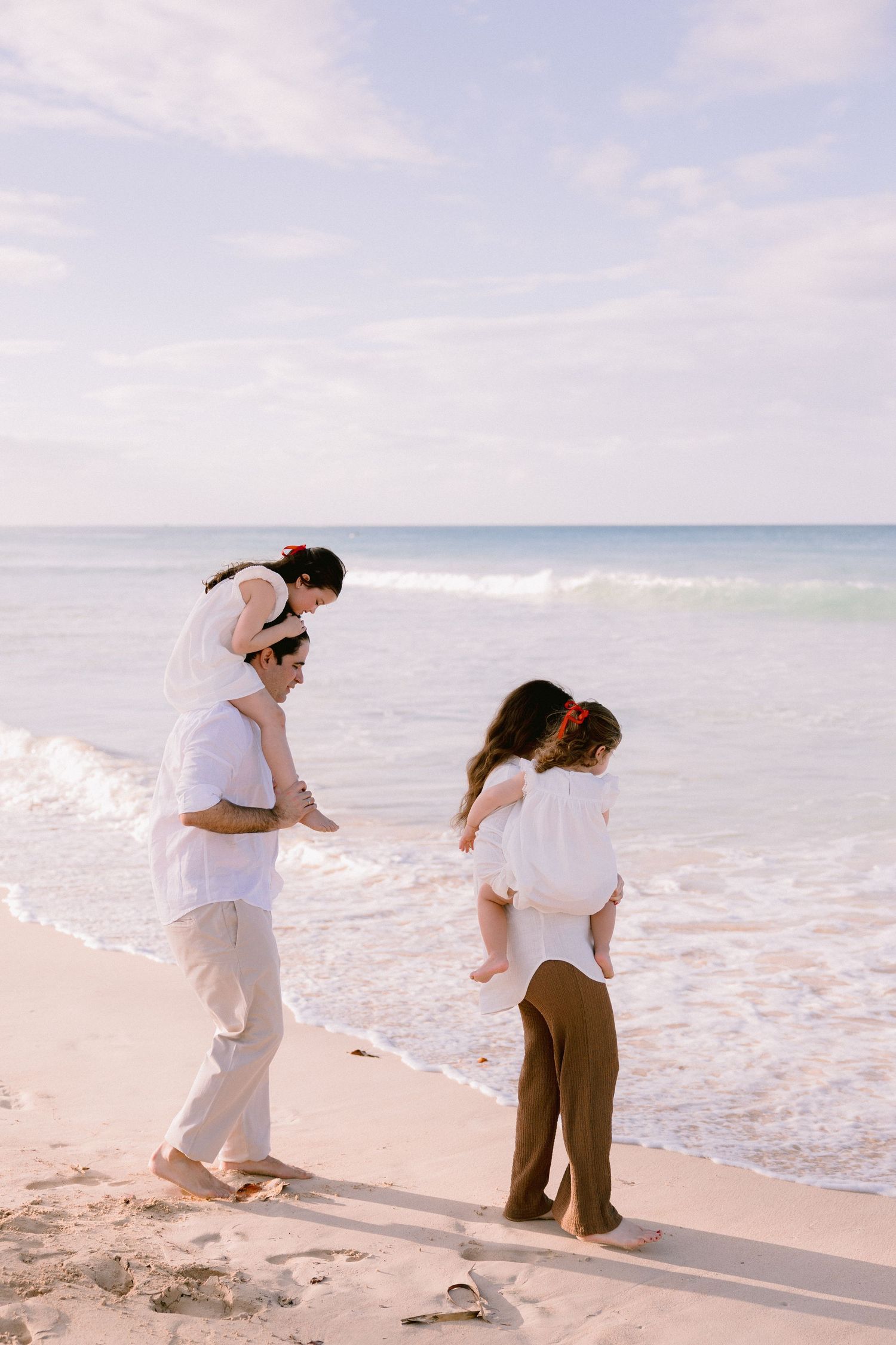 A couple in white and brown outfits dances together on a sunny beach with ocean waves in the background.
