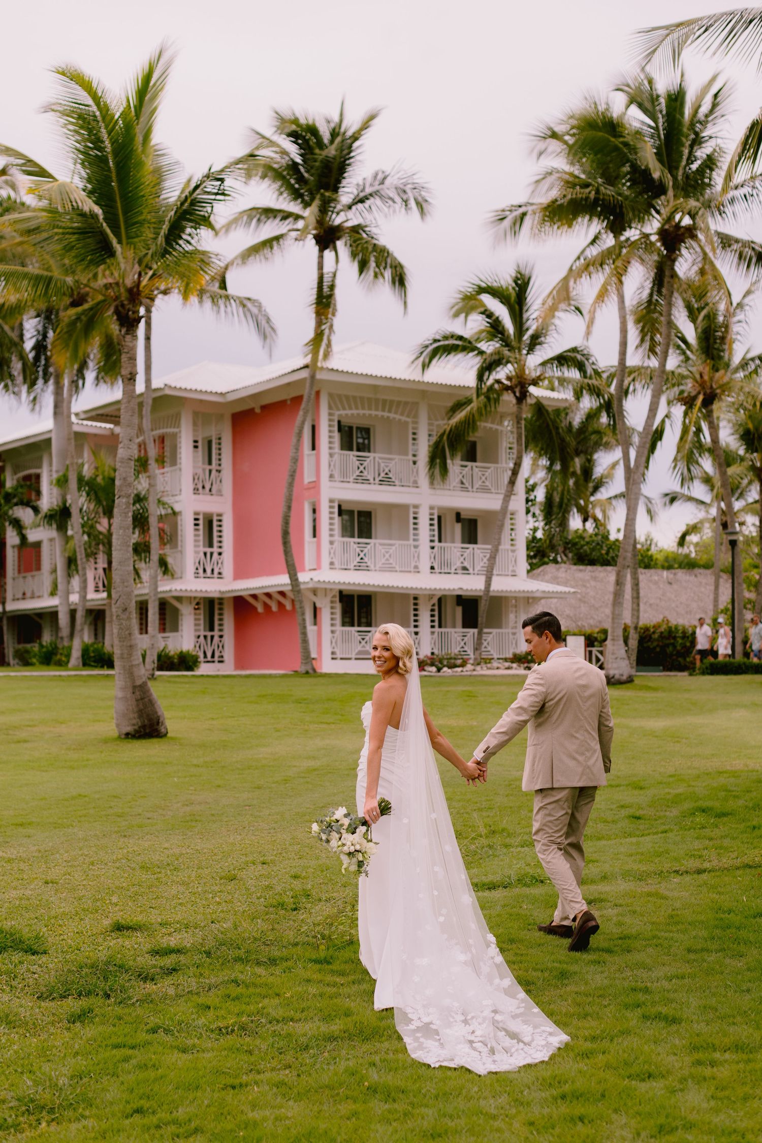 Bride and groom hold hands walking on tropical resort lawn with palm trees and pink building backdrop.