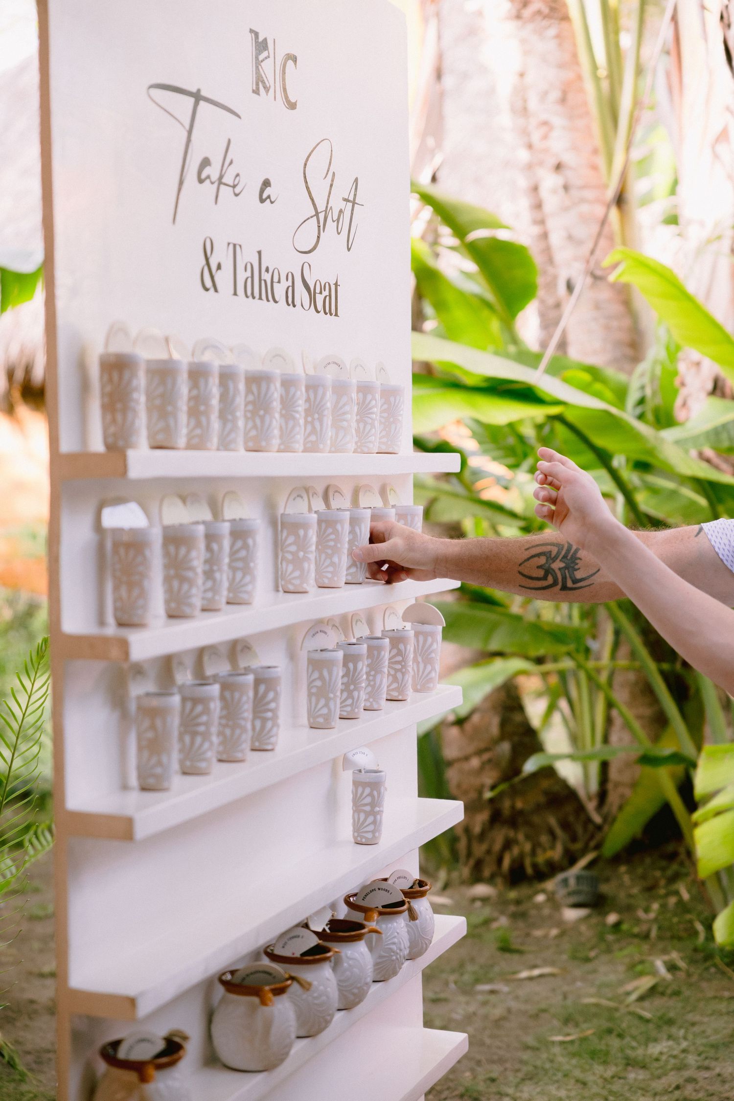 Wedding seating chart display with shot glasses on white shelves, reading Take a Shot and Take a Seat, outdoors.