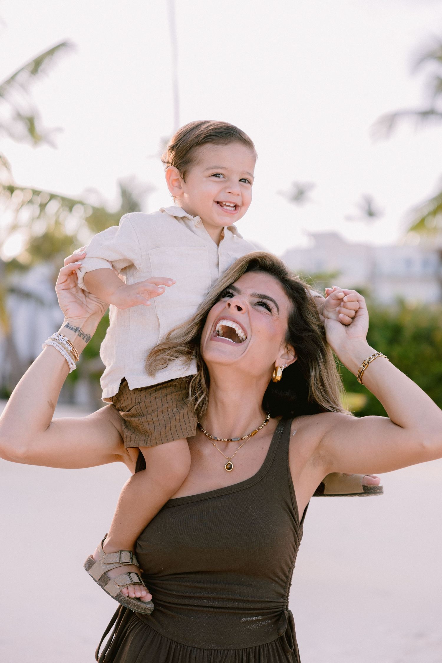 A woman in a brown top plays and laughs joyfully with a baby in white clothing outdoors on a sunny day.