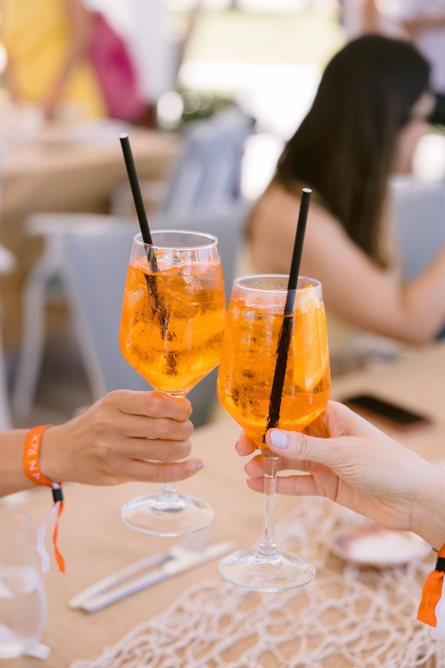 Two hands clinking glasses of orange Aperol spritz with black straws and orange slices at a sunny outdoor table.