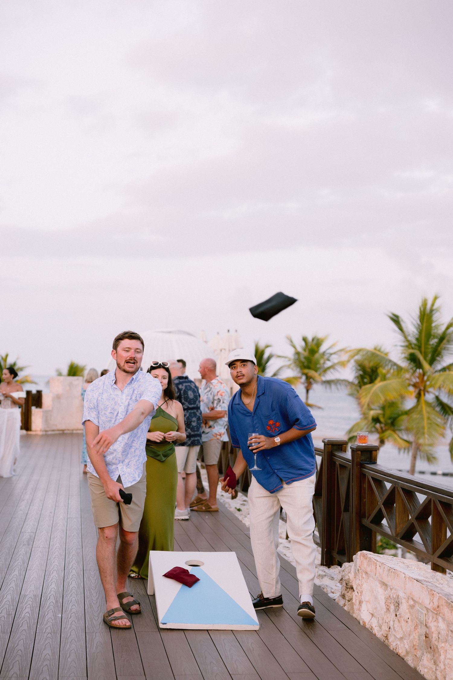 Guests in summer attire play cornhole on a wooden seaside deck at sunset while a bag flies through the air near palm trees.