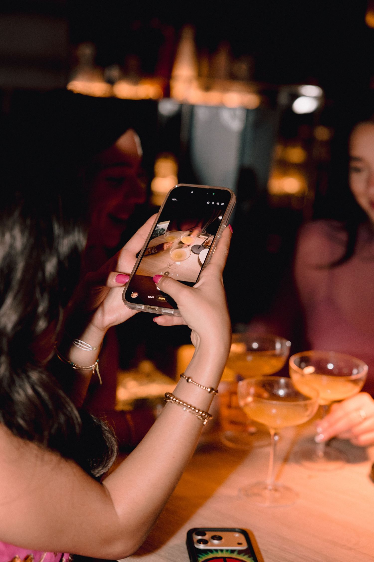 Woman with gold bracelets photographs two coupe glasses of pink cocktails on a bar table with her smartphone in a warmly lit lounge.