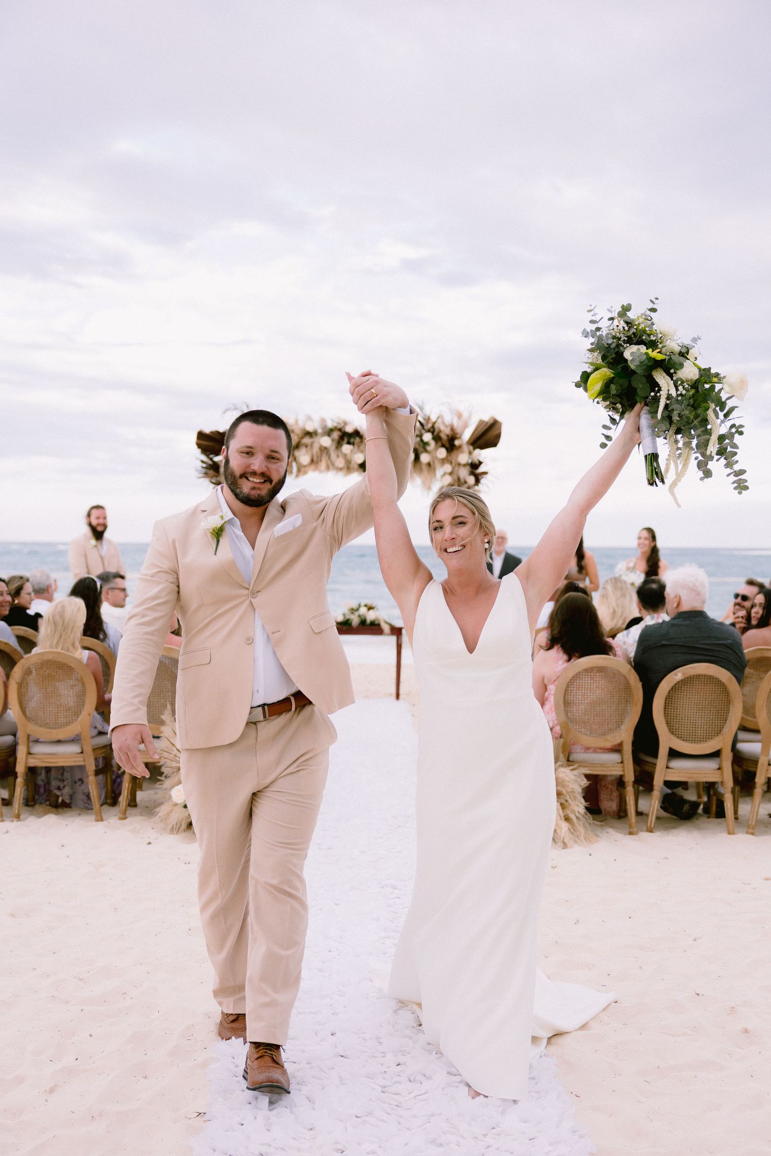 Joyful newlyweds celebrate on a beach ceremony, bride in white gown holding bouquet aloft, groom in tan suit.