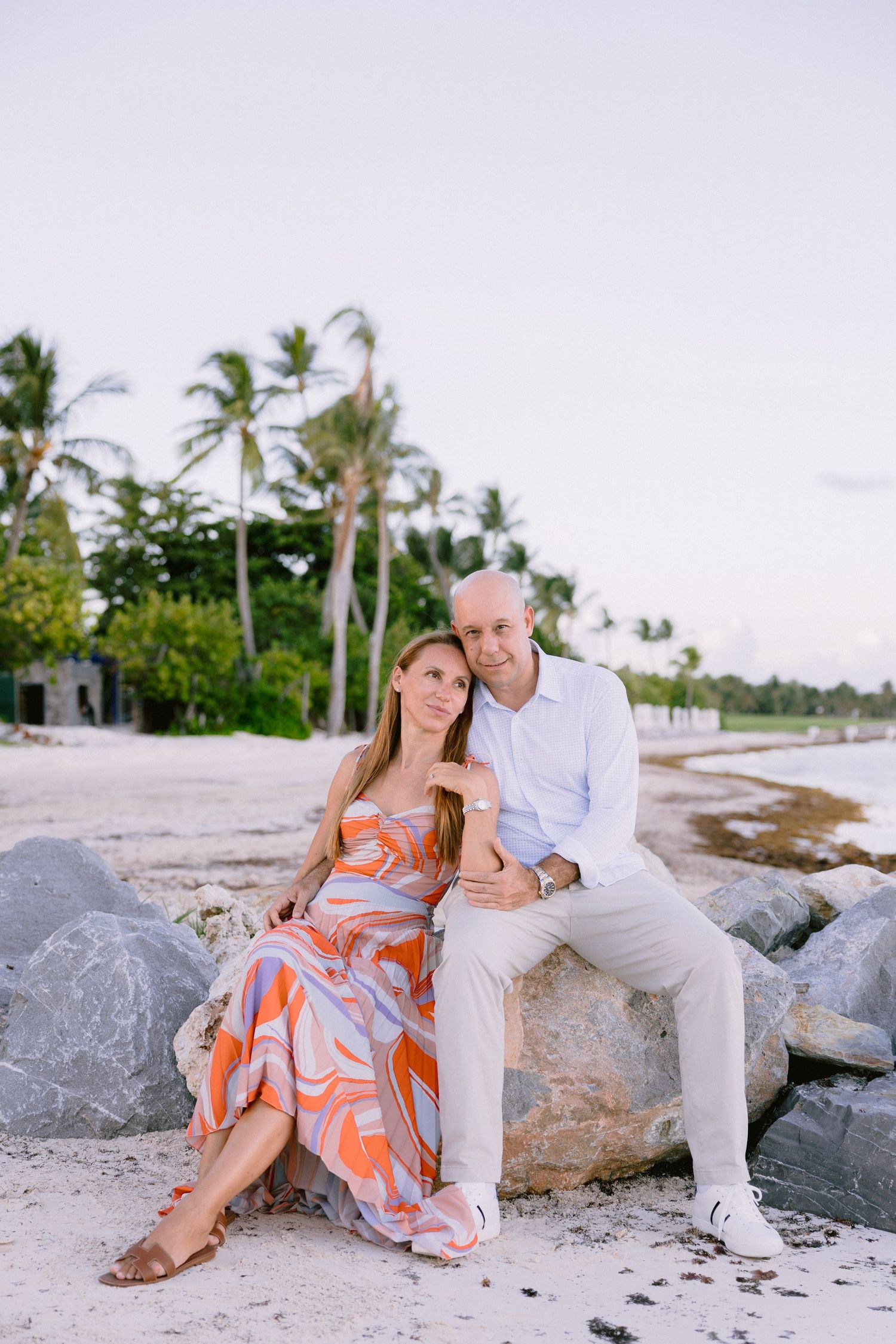 Couple sitting on rocks at a tropical beach, woman in orange dress, man in white shirt, palm trees background.