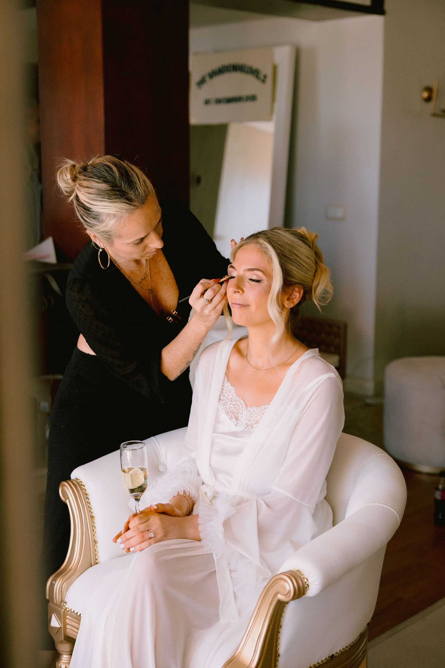 Makeup artist applying eye makeup to a bride in a white robe, seated in an elegant gold chair on her wedding day.