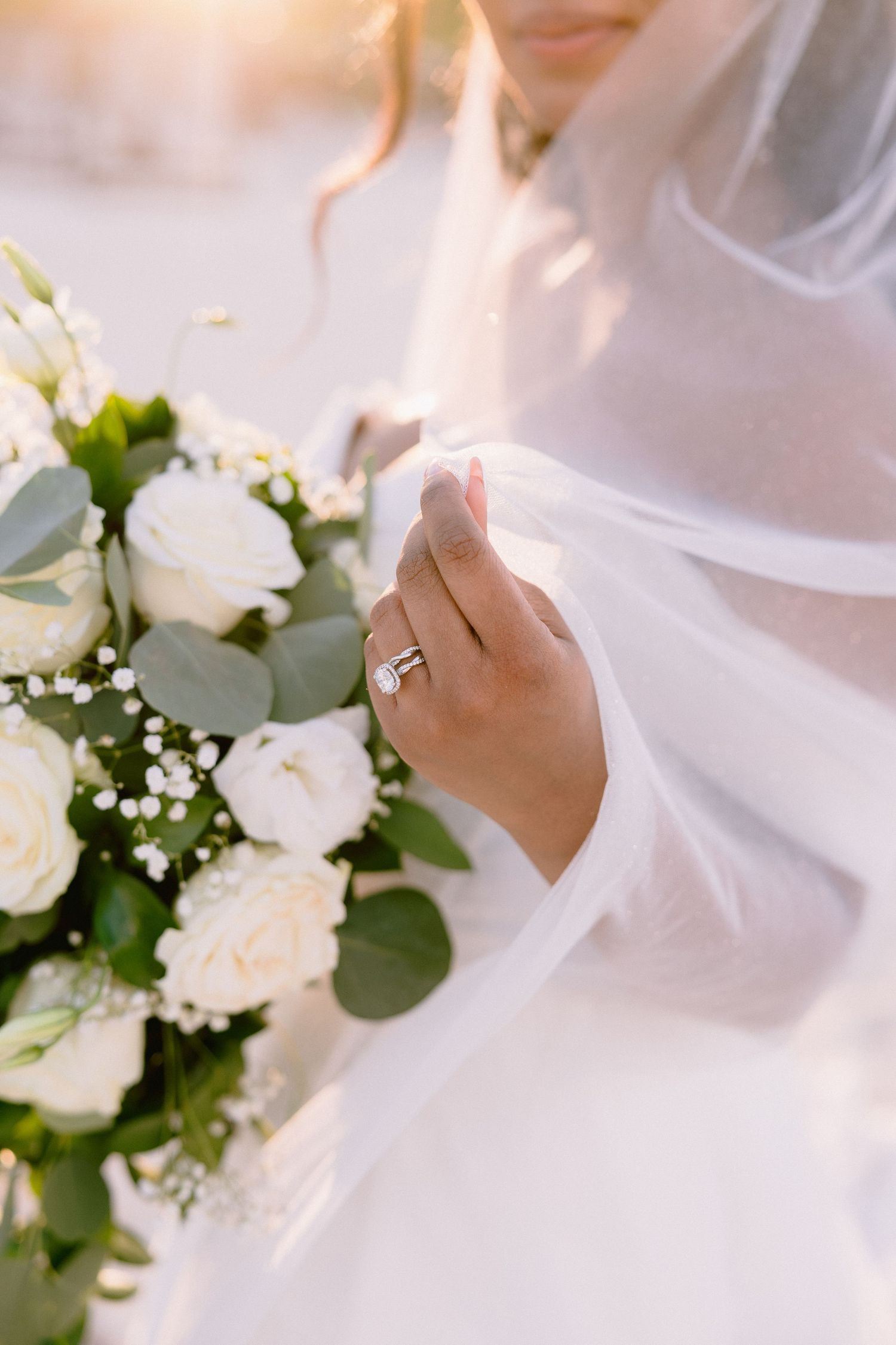 Bride holding white rose bouquet with eucalyptus, wearing diamond ring, veil draped over hand in golden sunlight.