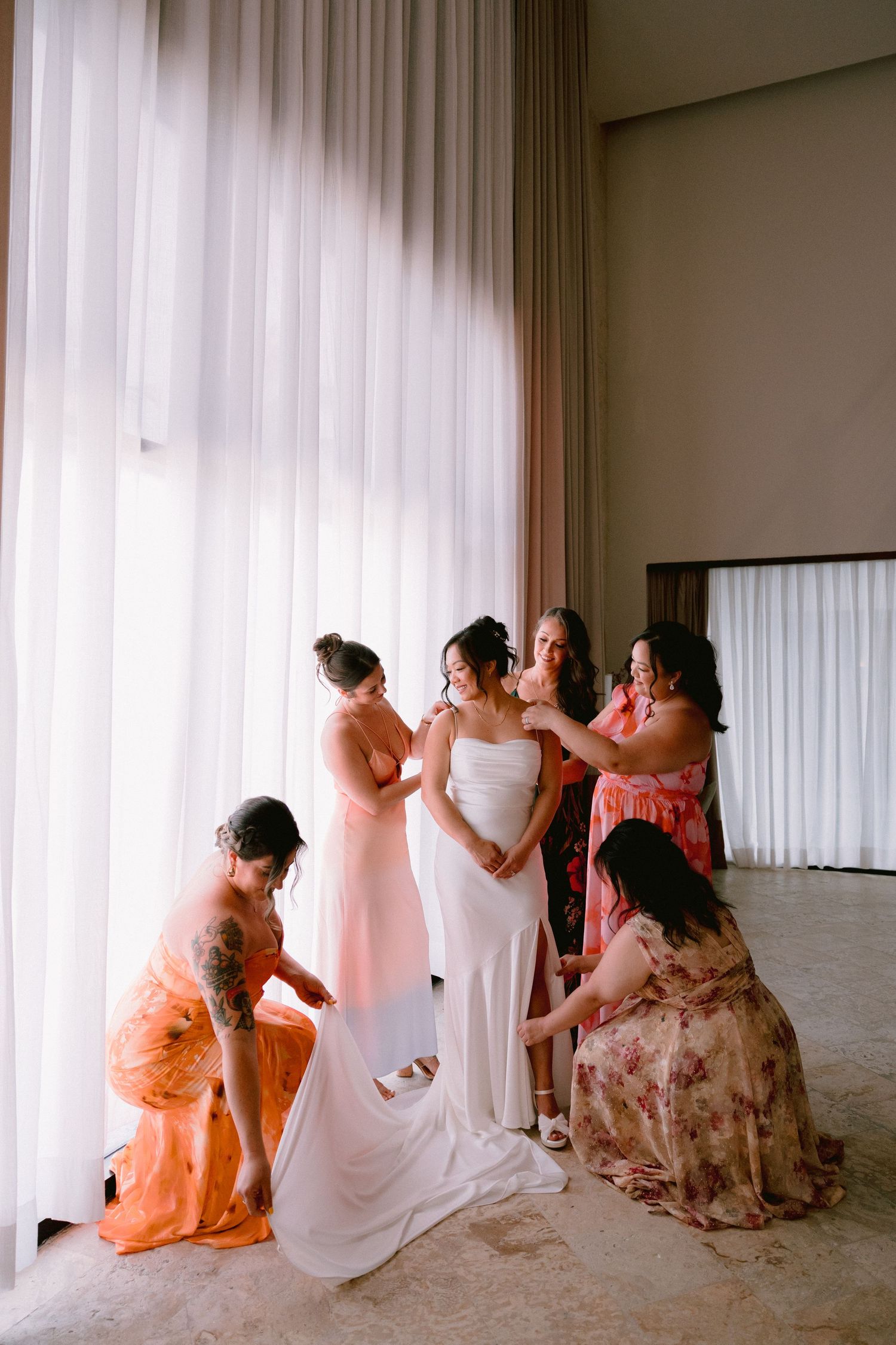 Bridesmaids in coral and floral dresses help a bride in a white gown prepare near sheer curtains.