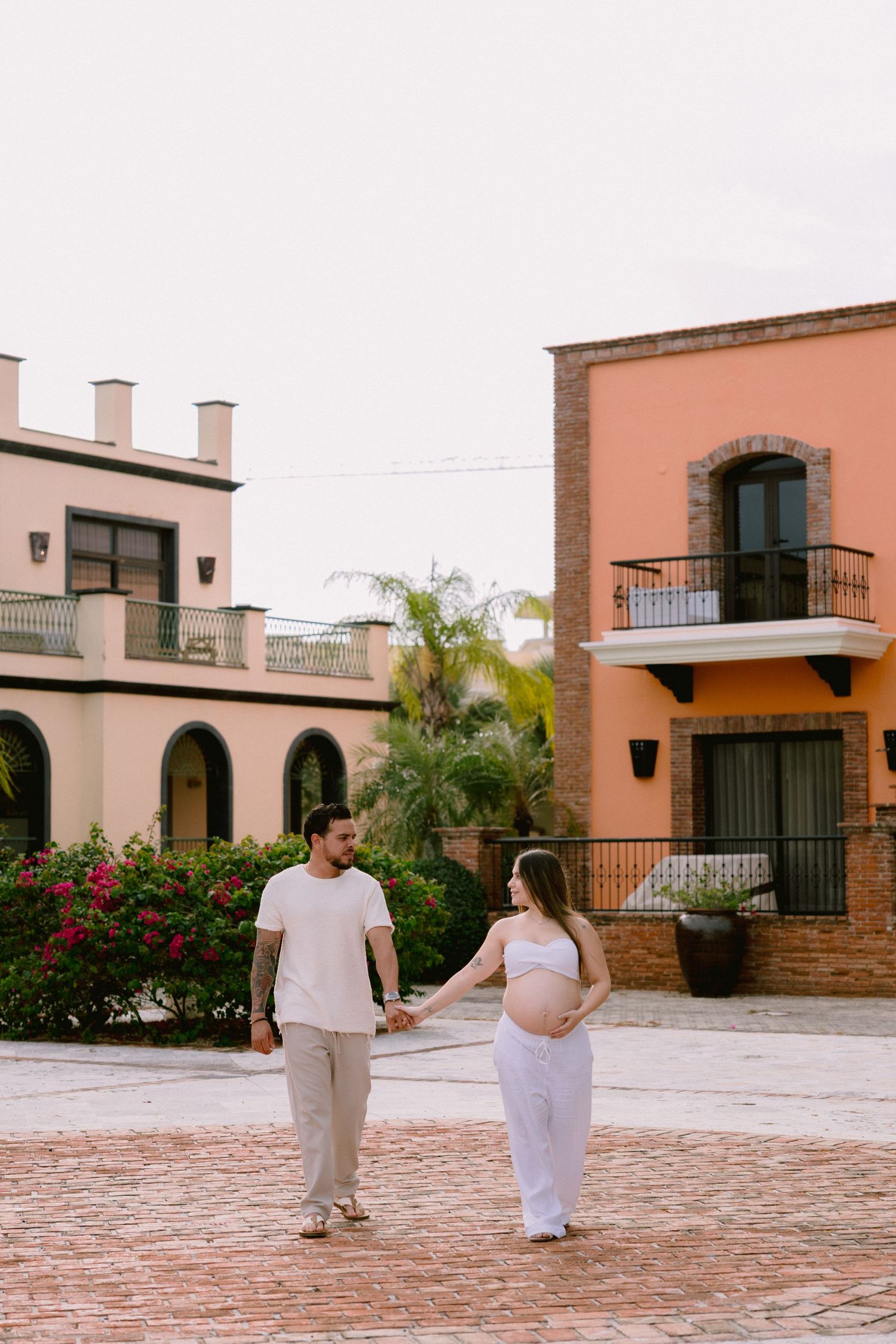 A couple walking hand in hand on a brick courtyard surrounded by Spanish colonial style architecture and vibrant flowers.