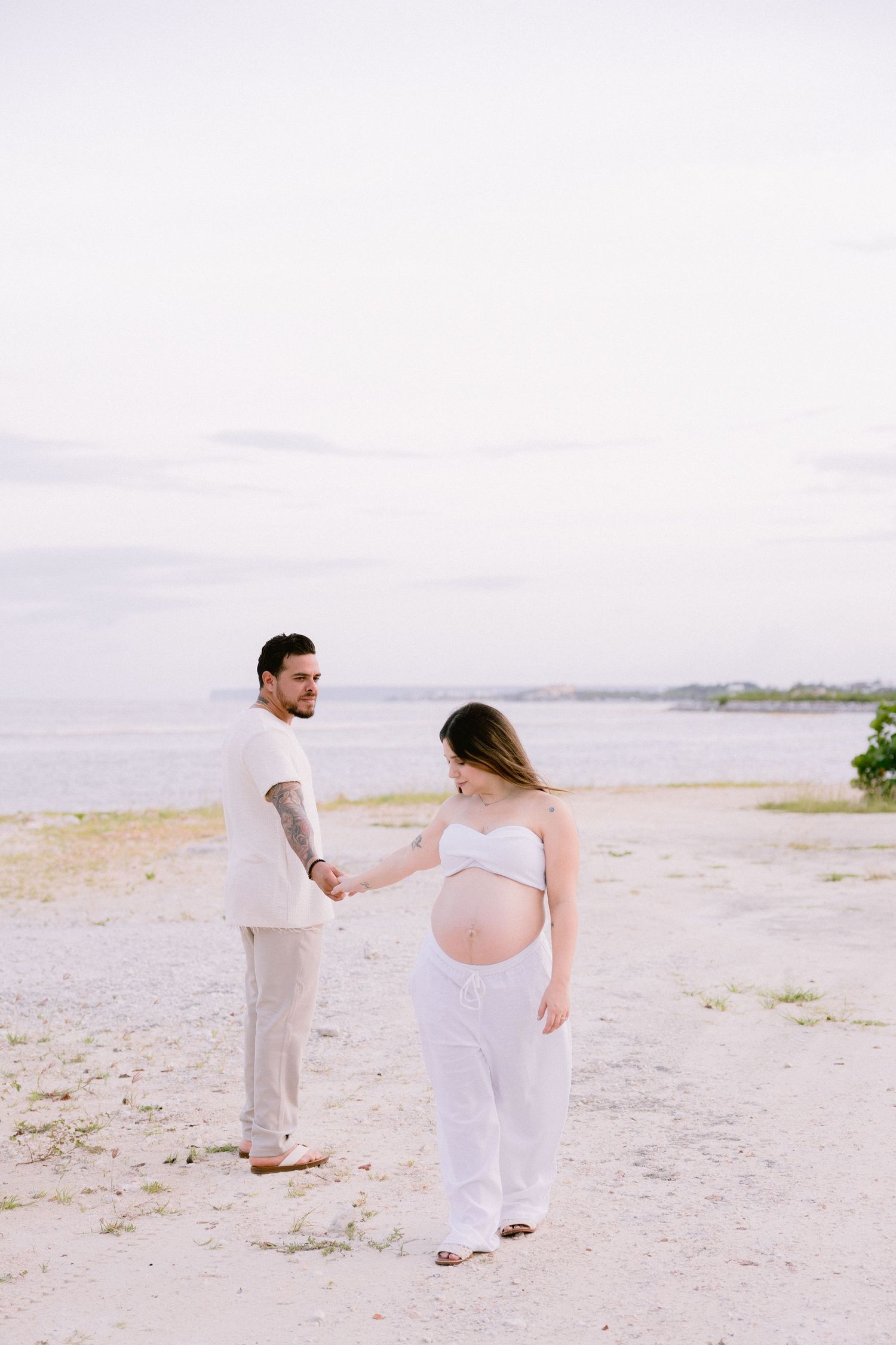 A couple in white stands on a sandy beach against an overcast sky during a maternity photoshoot.