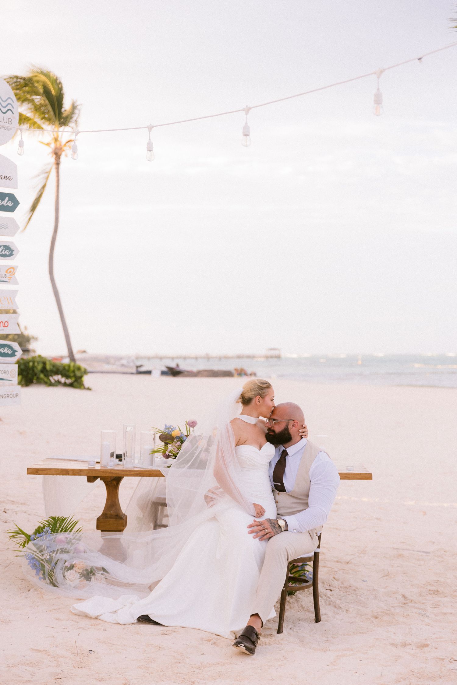 Bride and groom share a kiss at a romantic beach wedding table setup with string lights and a palm tree backdrop.