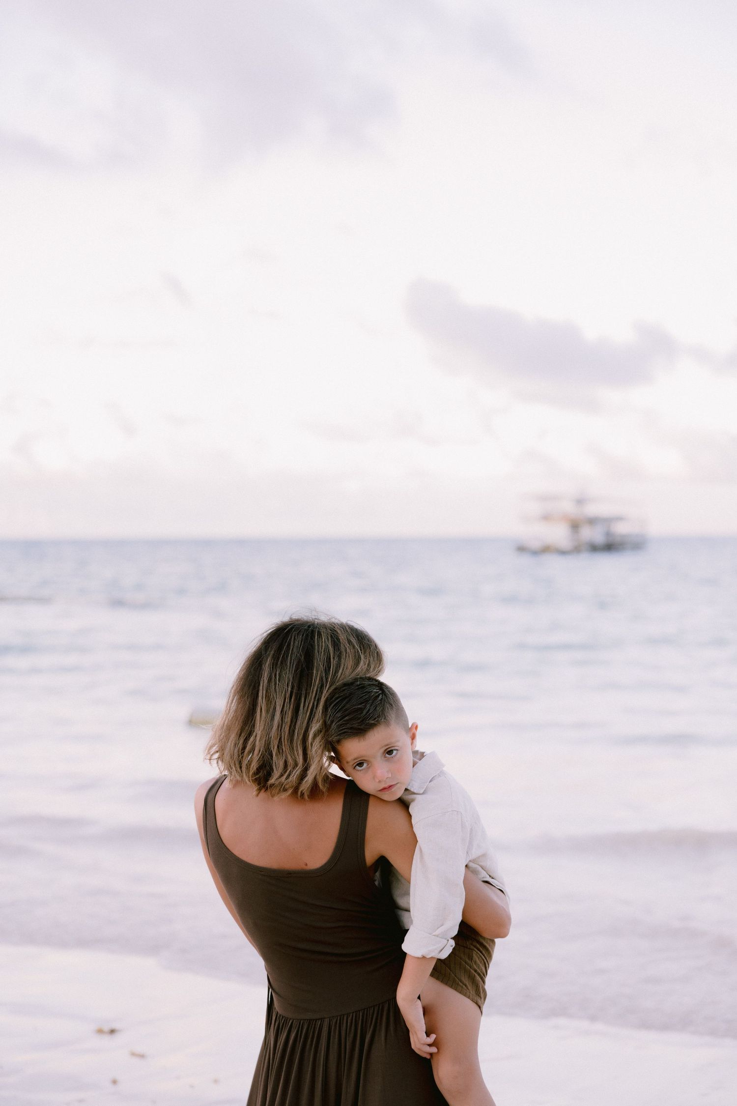 A parent in a brown dress holds a child while standing on a beach with a boat visible in the distance.