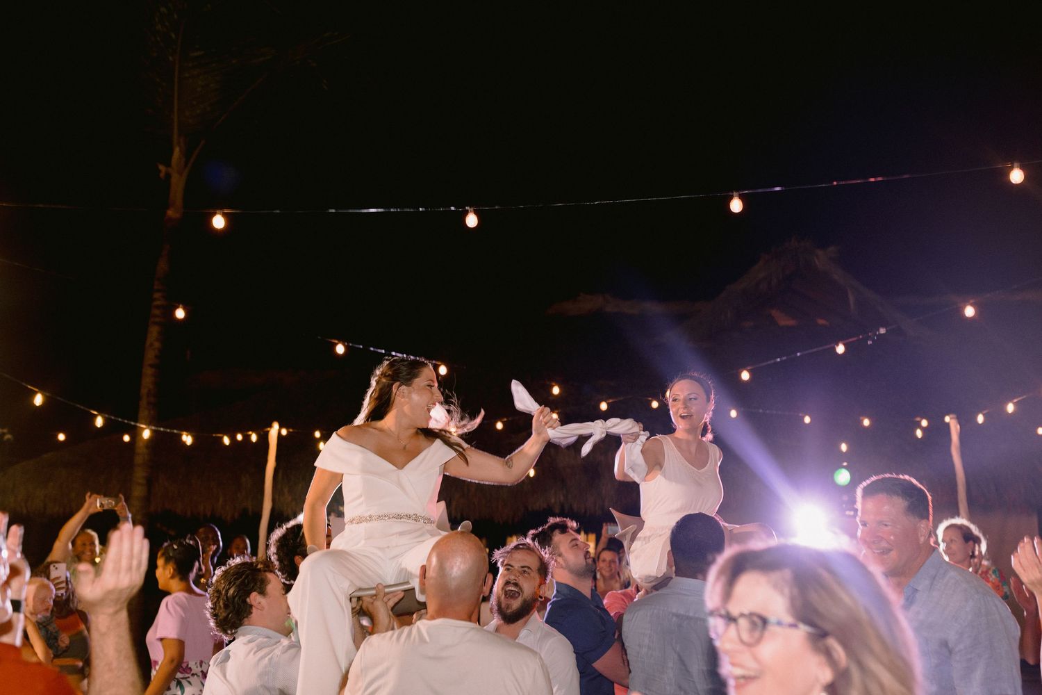 Bride and groom lifted on chairs during lively outdoor wedding reception at night with string lights.